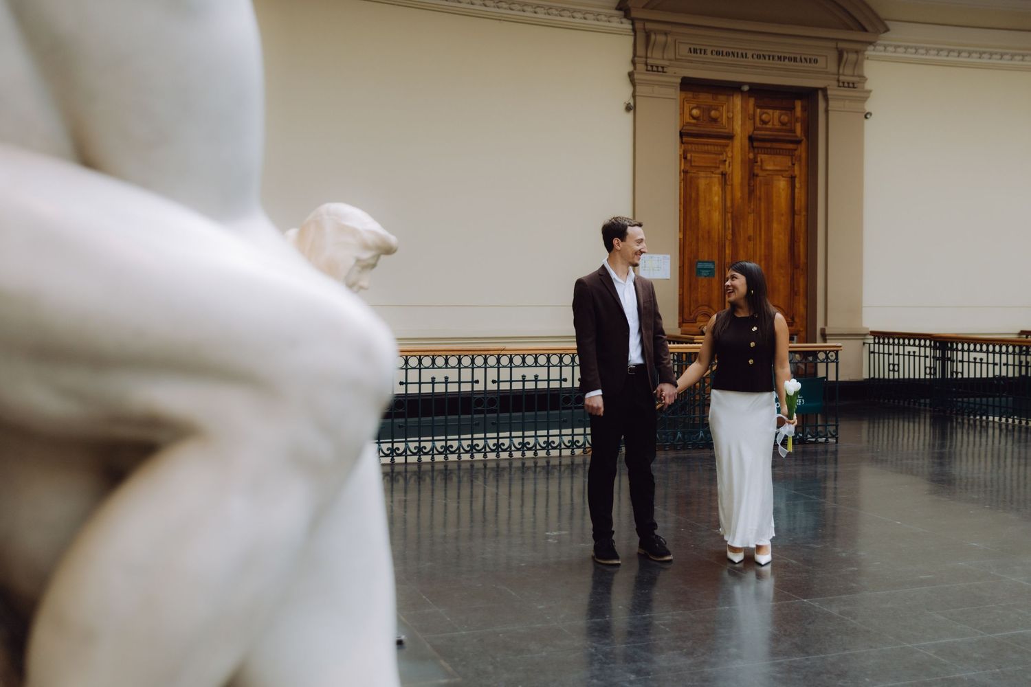Couple holding hands in a grand museum hall with marble sculptures and ornate wooden doors behind them.