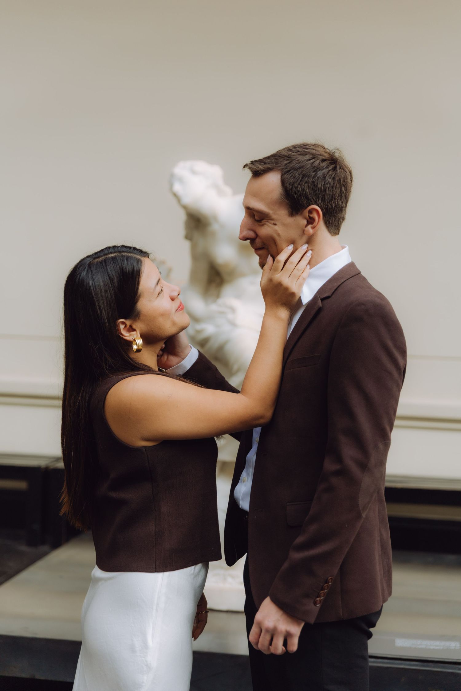 Woman tenderly touching mans face as they smile at each other with a classical marble statue behind them.