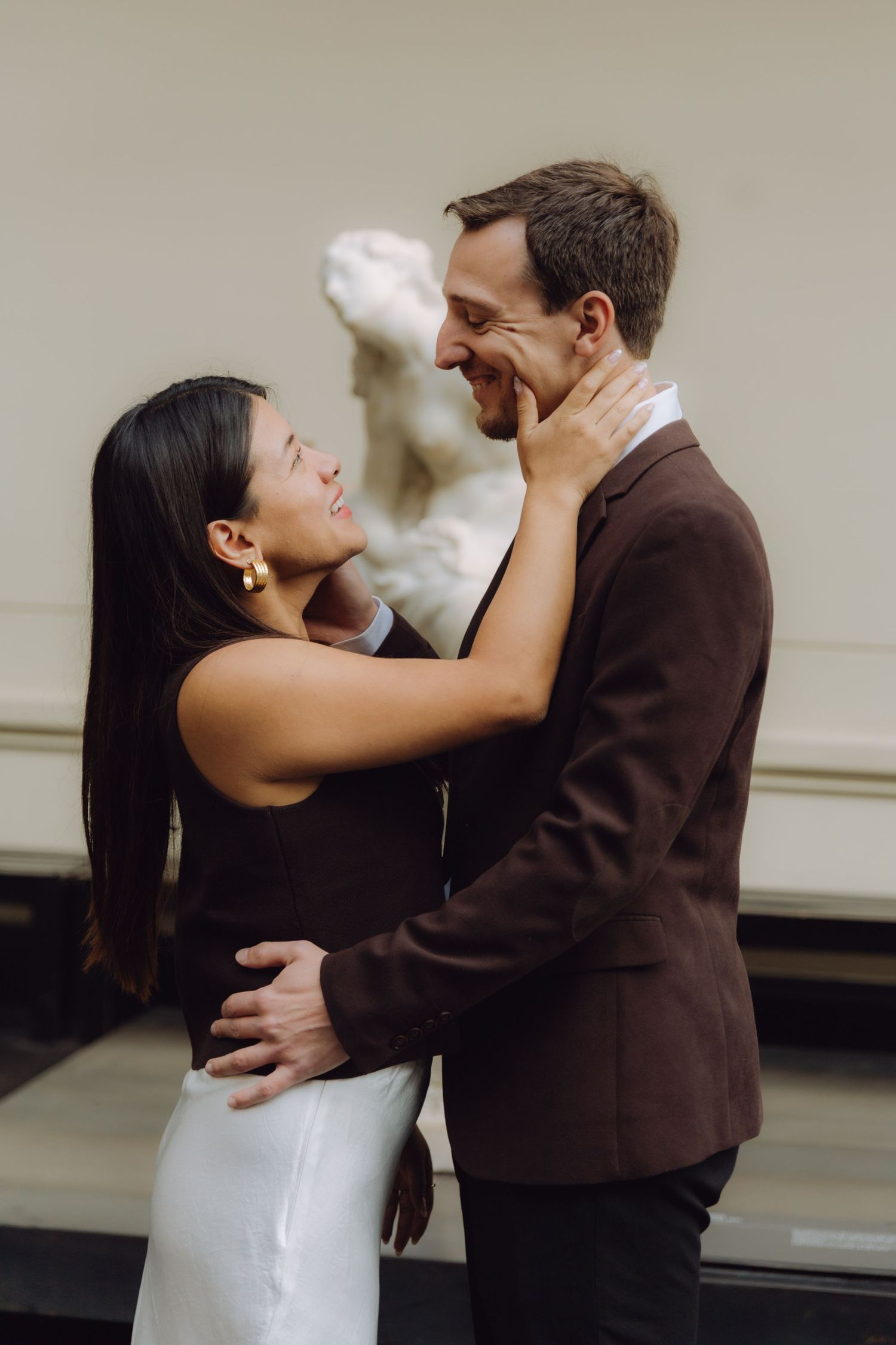 Couple embracing and smiling at each other in a museum gallery with a white marble sculpture backdrop.