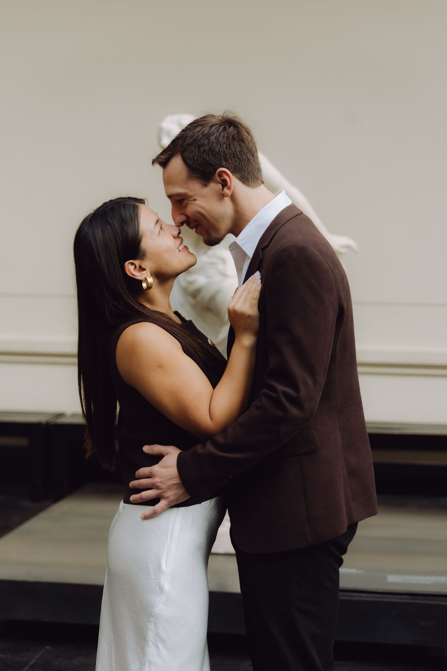 Couple sharing an intimate moment, nearly kissing, in a softly lit museum gallery with sculptures nearby.