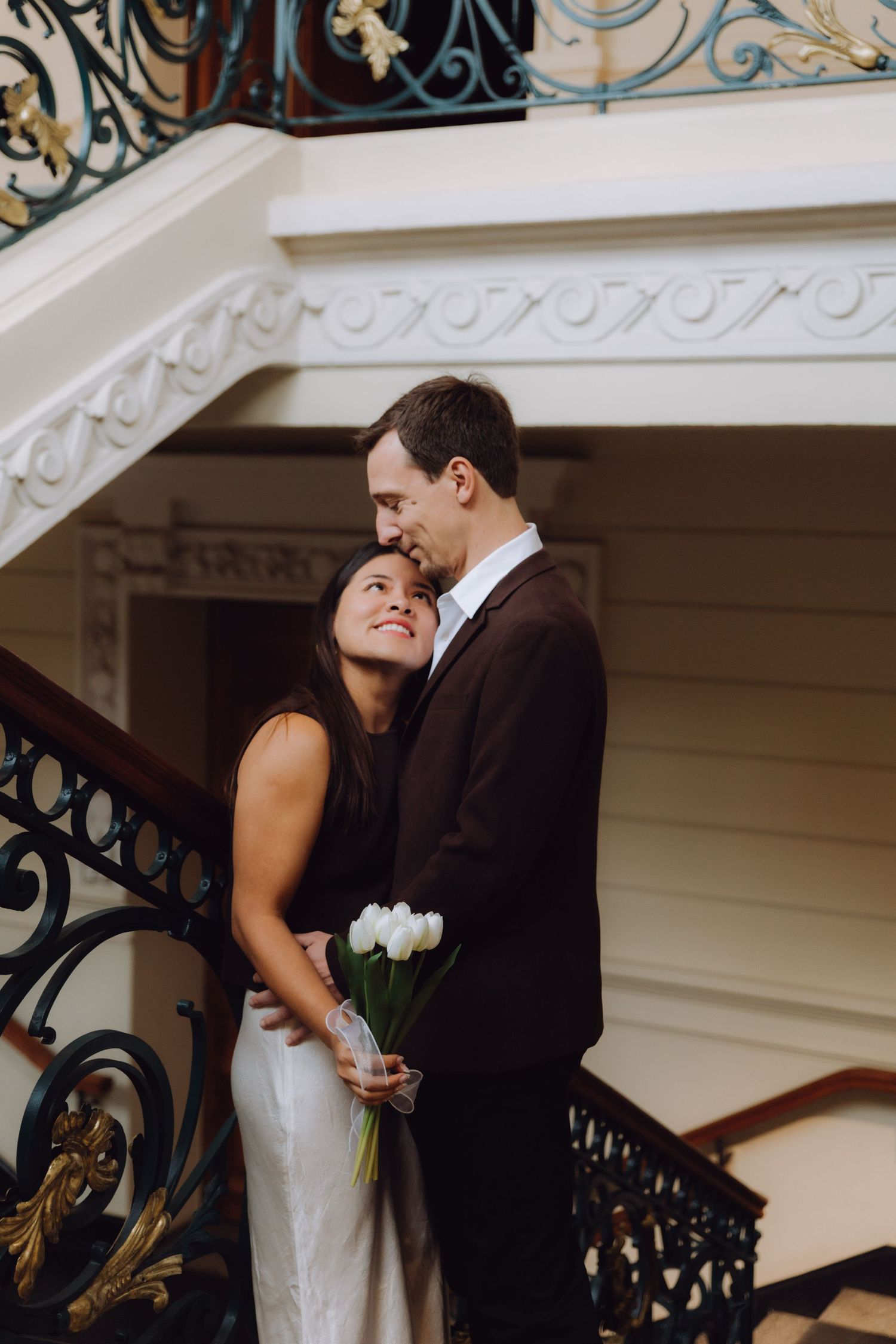 Couple embracing on an ornate staircase, woman holding white tulips and smiling up at her partner.
