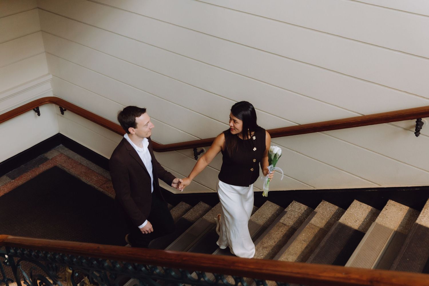 Couple holding hands while walking up wooden stairs, woman carrying white tulips, viewed from above.