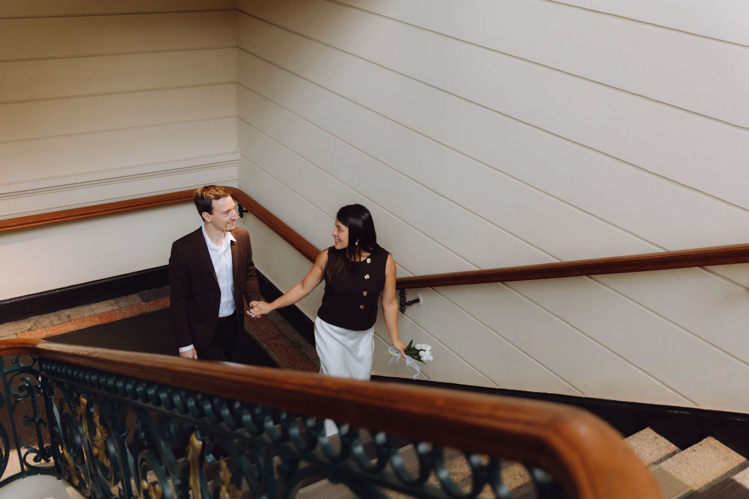 Couple holding hands walking up elegant staircase with ornate iron railings in a grand historic building.