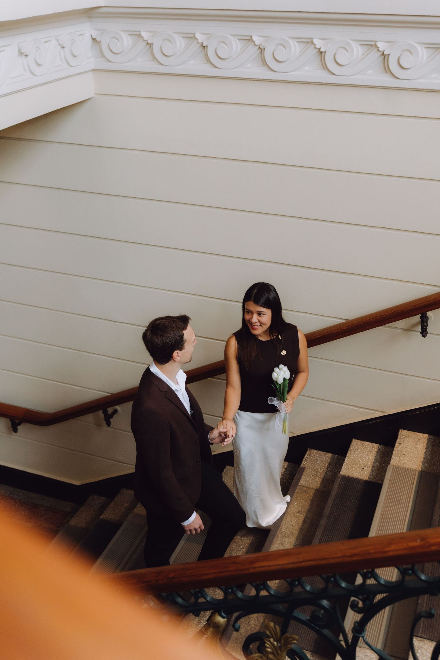 Bride holding white bouquet and groom holding hands on ornate staircase in a classical building.