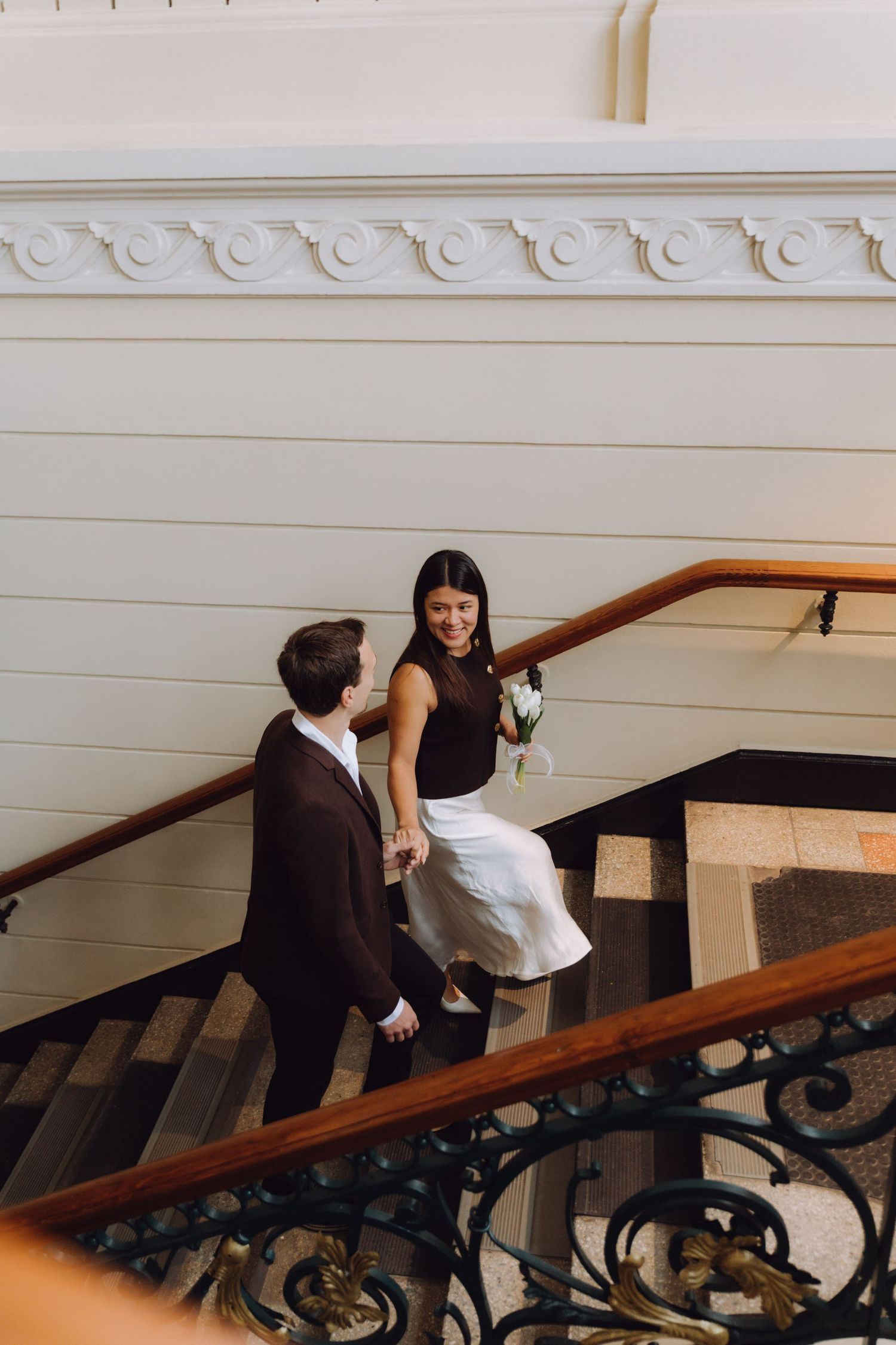 Smiling bride with white bouquet and groom walking up grand staircase with decorative iron railings.
