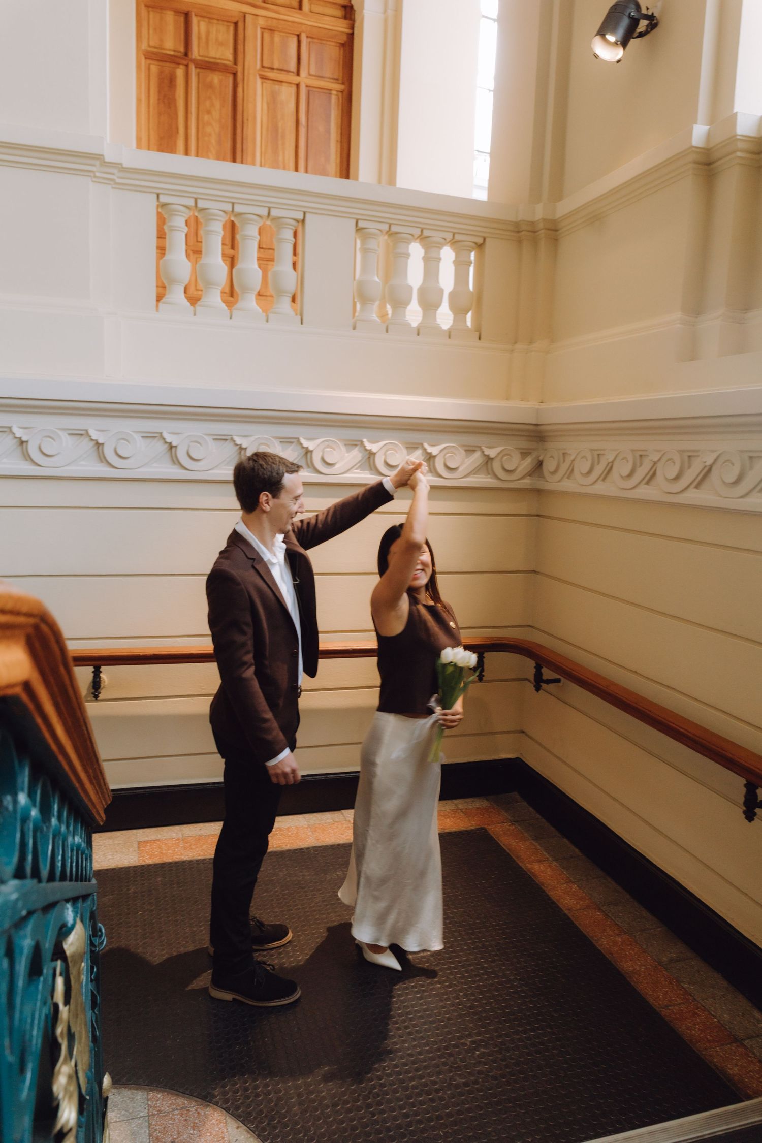 Groom spinning bride in white skirt on landing of elegant historic building staircase.