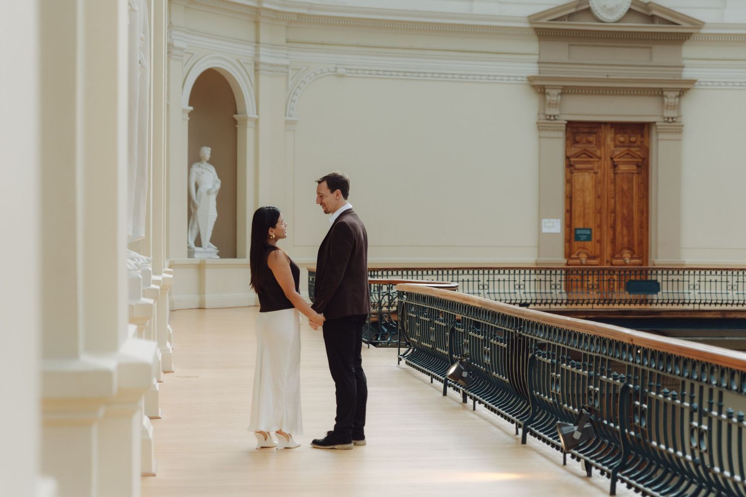 Couple holding hands facing each other on balcony of neoclassical museum with marble statues.