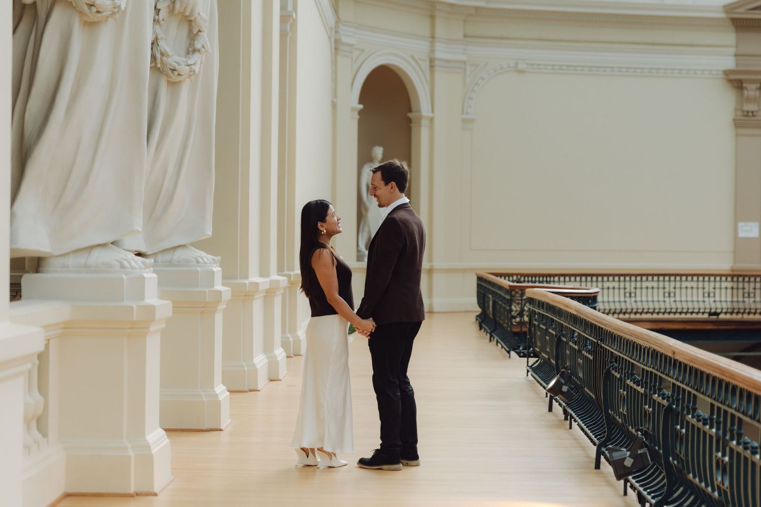 Elegant couple holding hands and gazing at each other beside grand columns in a classical hall.