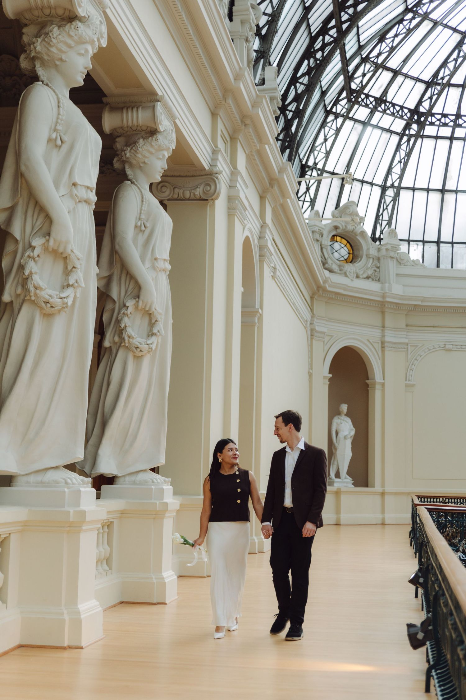 Couple walking hand in hand past towering white marble statues in ornate museum with glass ceiling.