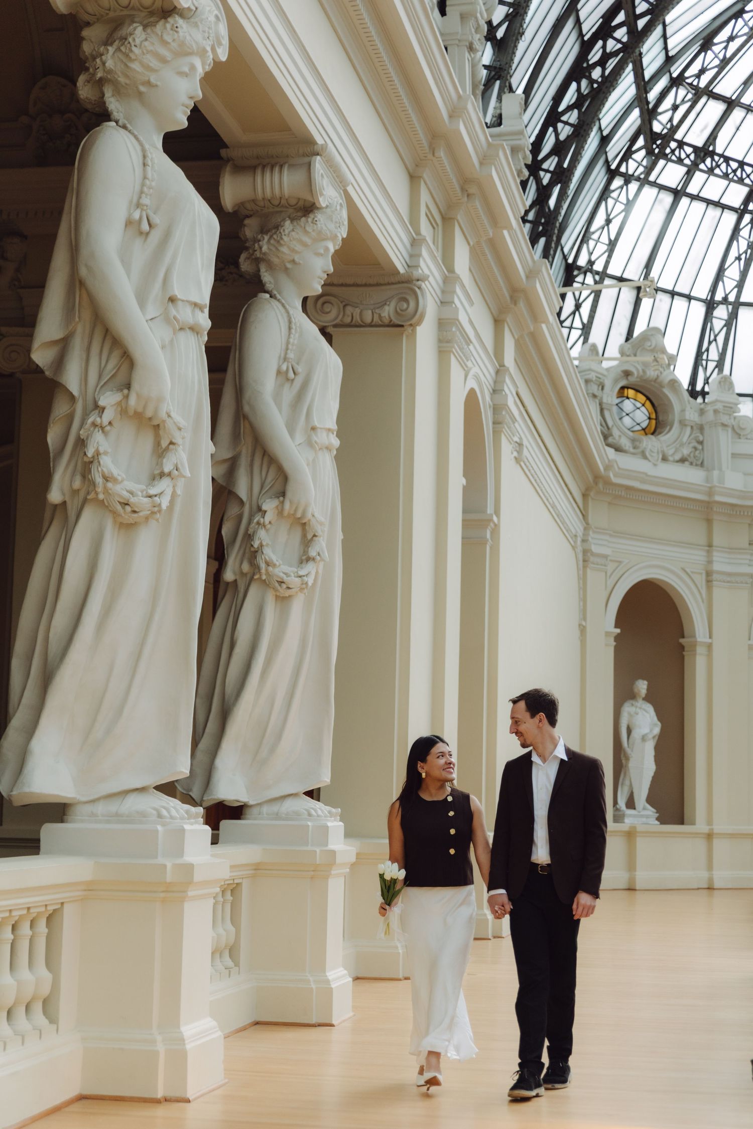 Couple strolling past classical marble caryatid statues in grand museum hall with glass skylight.