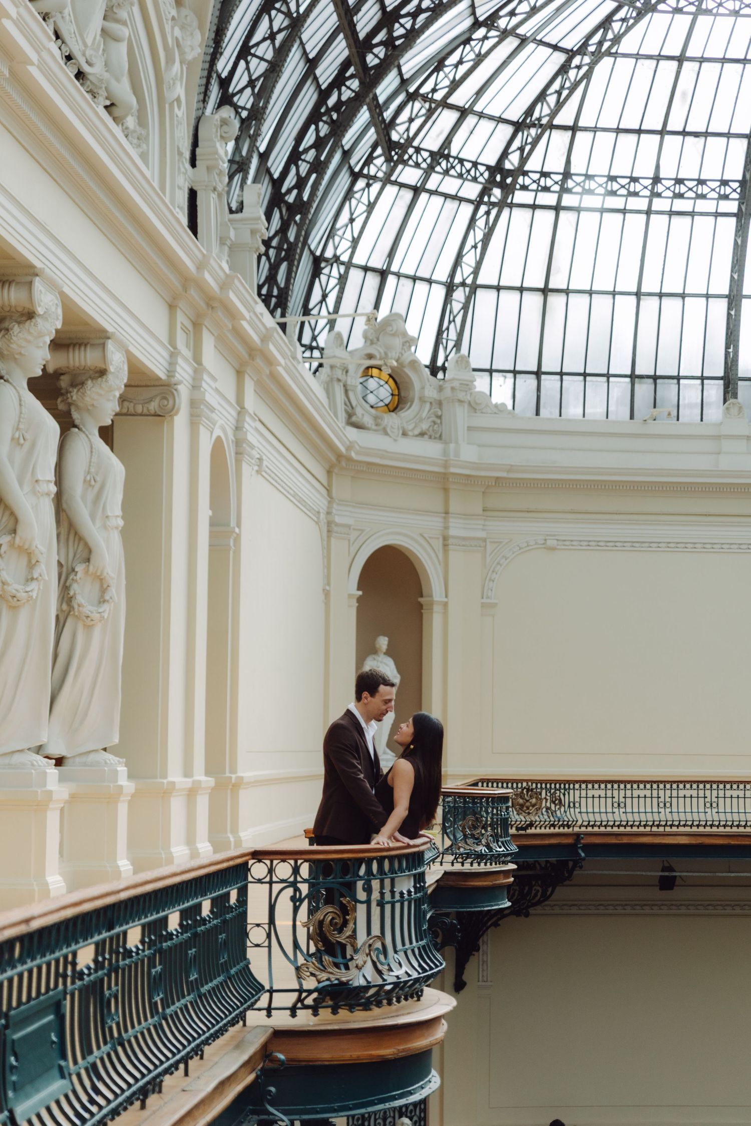 Couple sharing romantic moment on ornate balcony beneath stunning glass dome in historic building.