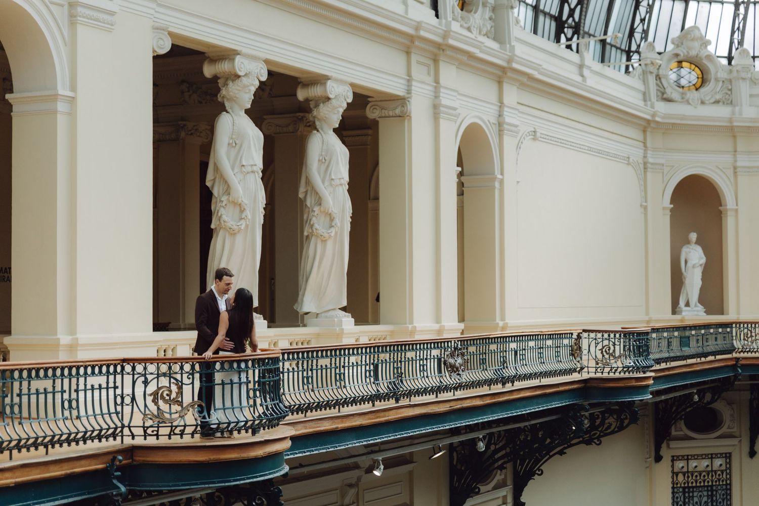 Couple standing on decorative iron balcony surrounded by classical marble statues in grand museum.