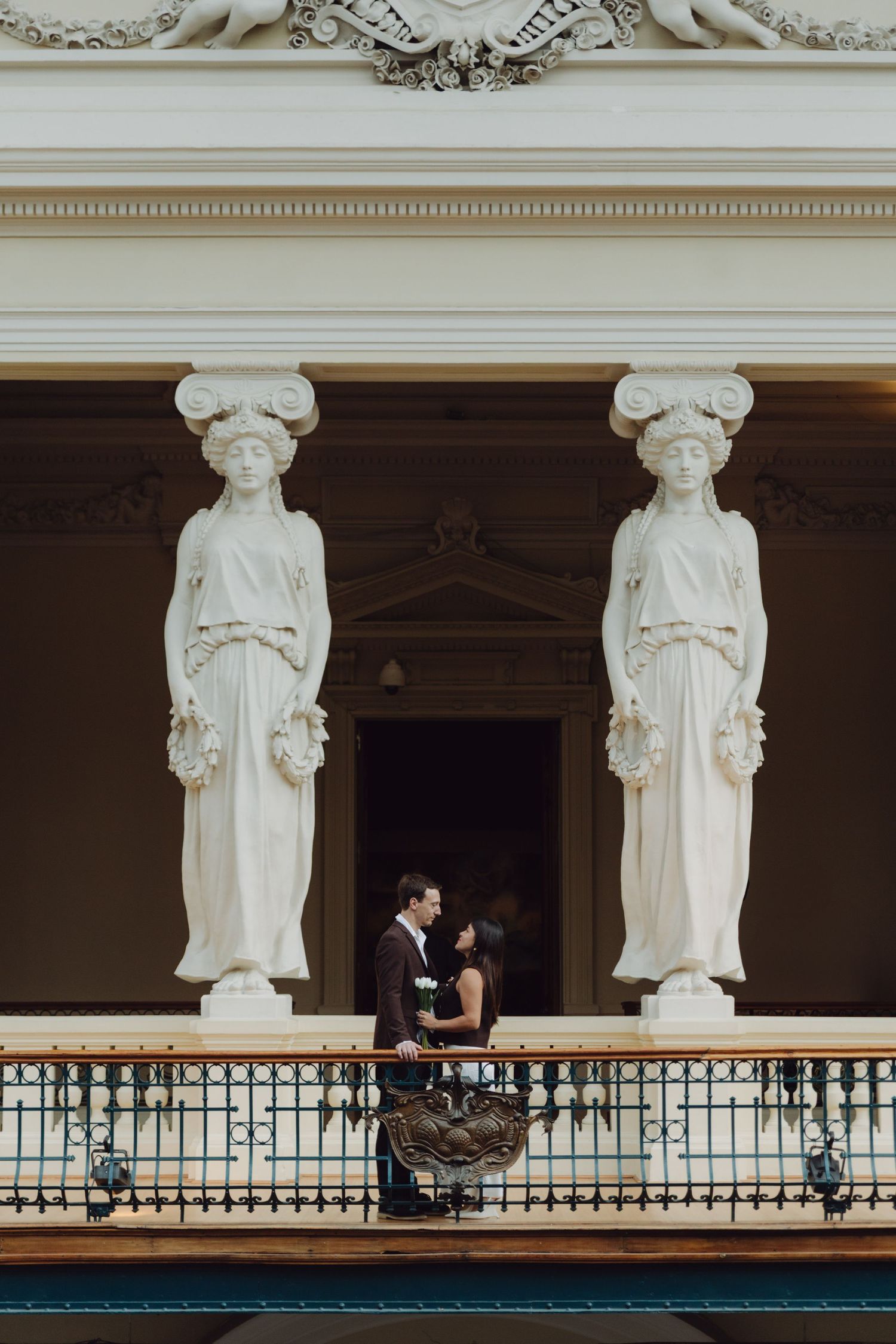 Couple embracing on ornate balcony flanked by classical caryatid statues at elegant neoclassical building.