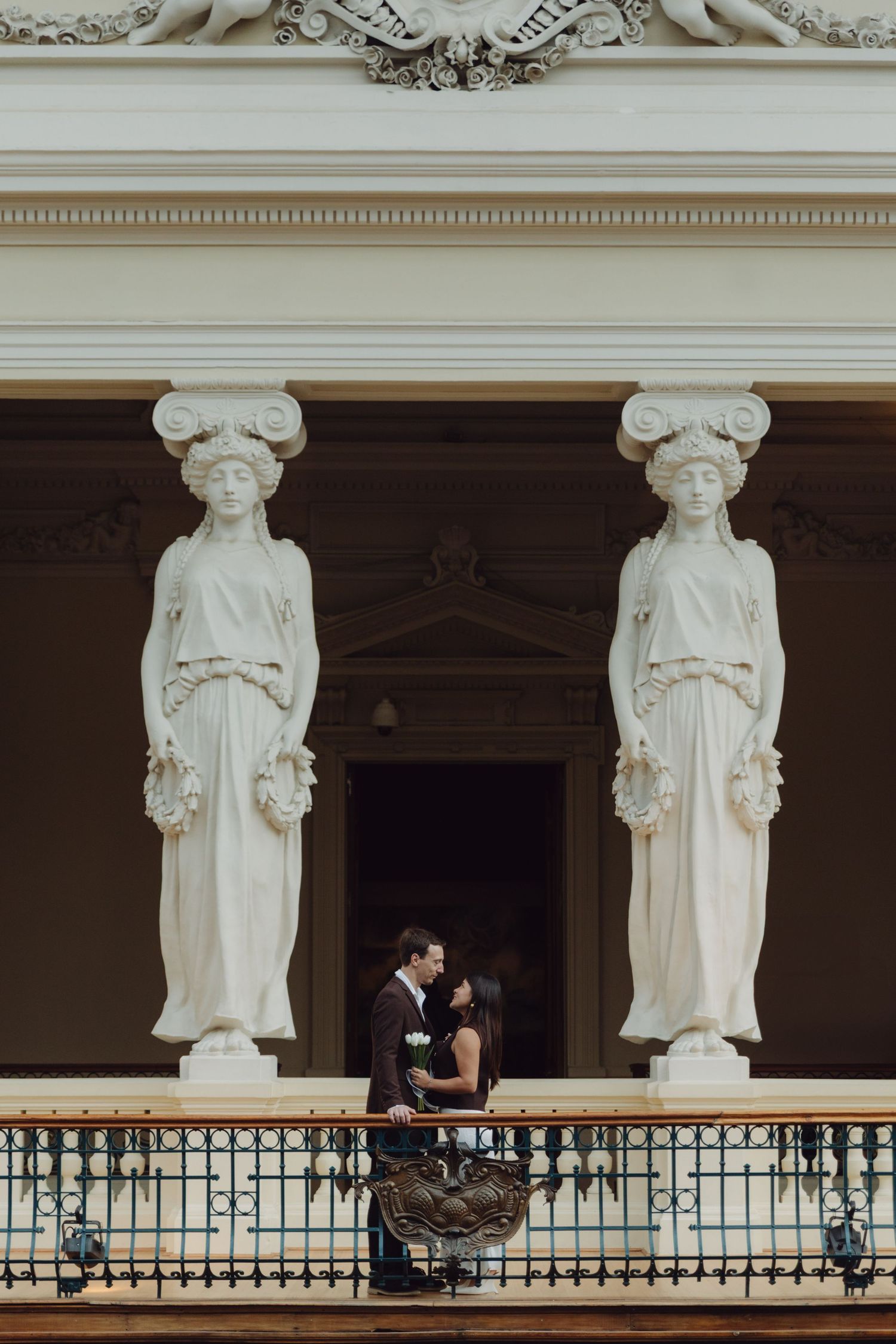 Romantic couple kissing between two white caryatid columns on decorative balcony of historic building.