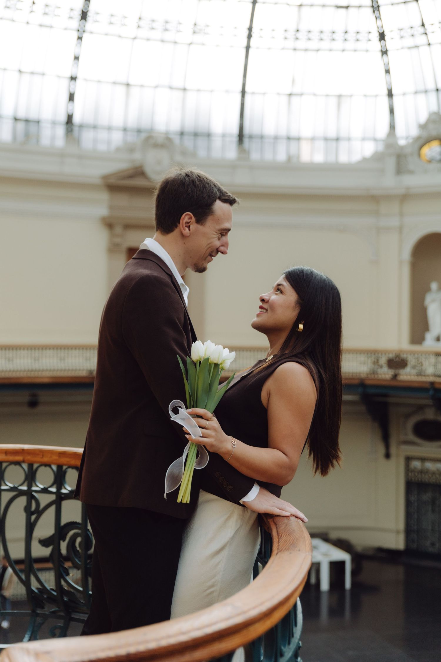 Smiling couple holding white tulip bouquet inside grand museum with glass dome ceiling and balconies.