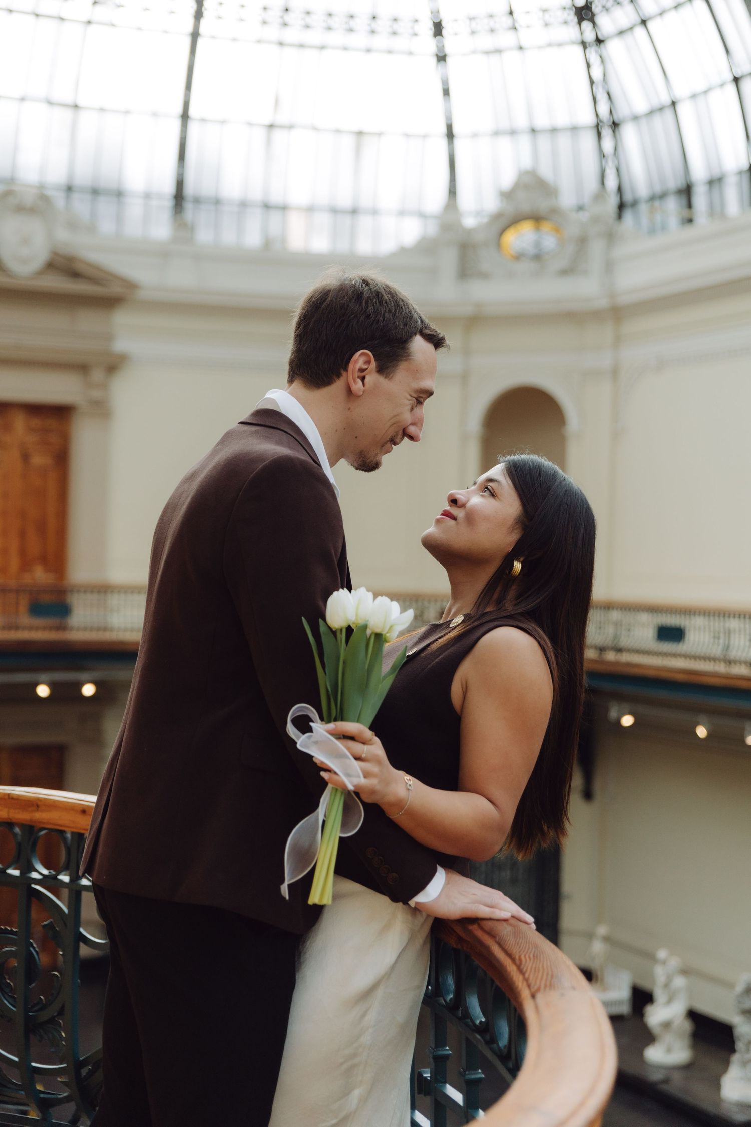Elegant couple sharing intimate moment with white tulips under stunning glass rotunda of classic building.