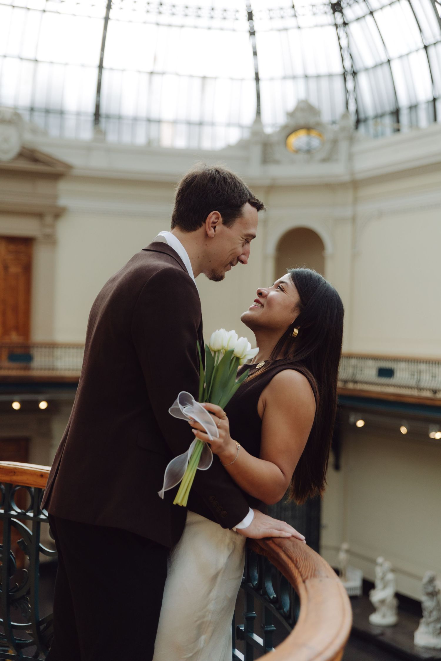 Couple about to kiss holding white tulip bouquet beneath ornate glass dome in historic interior.