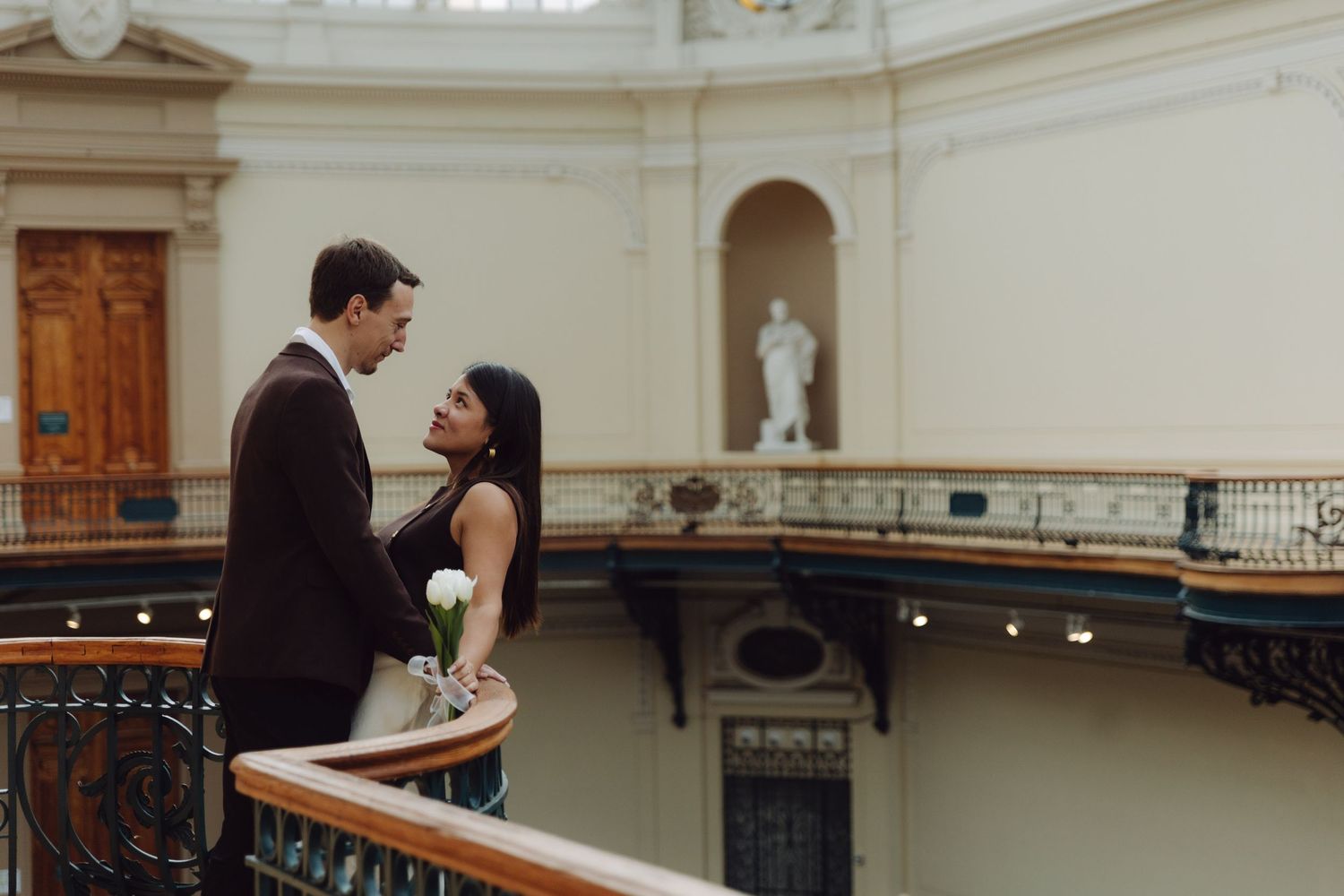 Stylish couple gazing at each other on curved balcony railing inside classical building with statue.