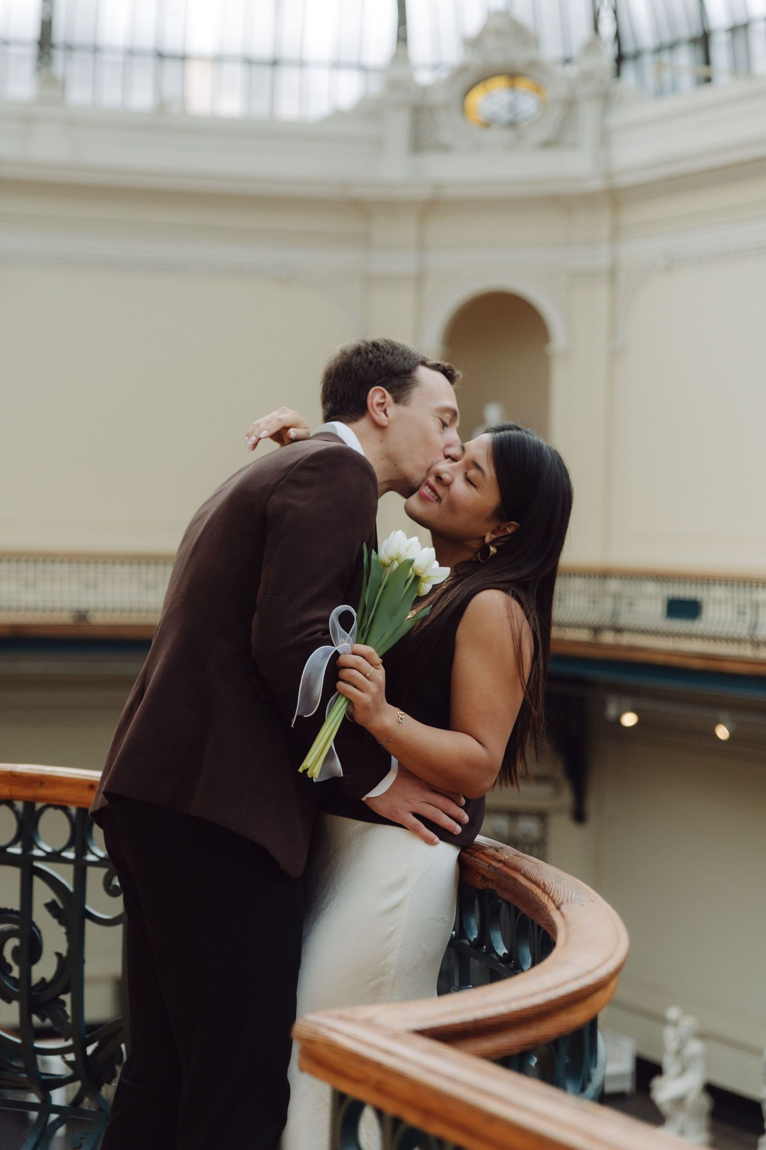 Couple sharing tender kiss while holding white tulip bouquet on sweeping staircase of grand building.