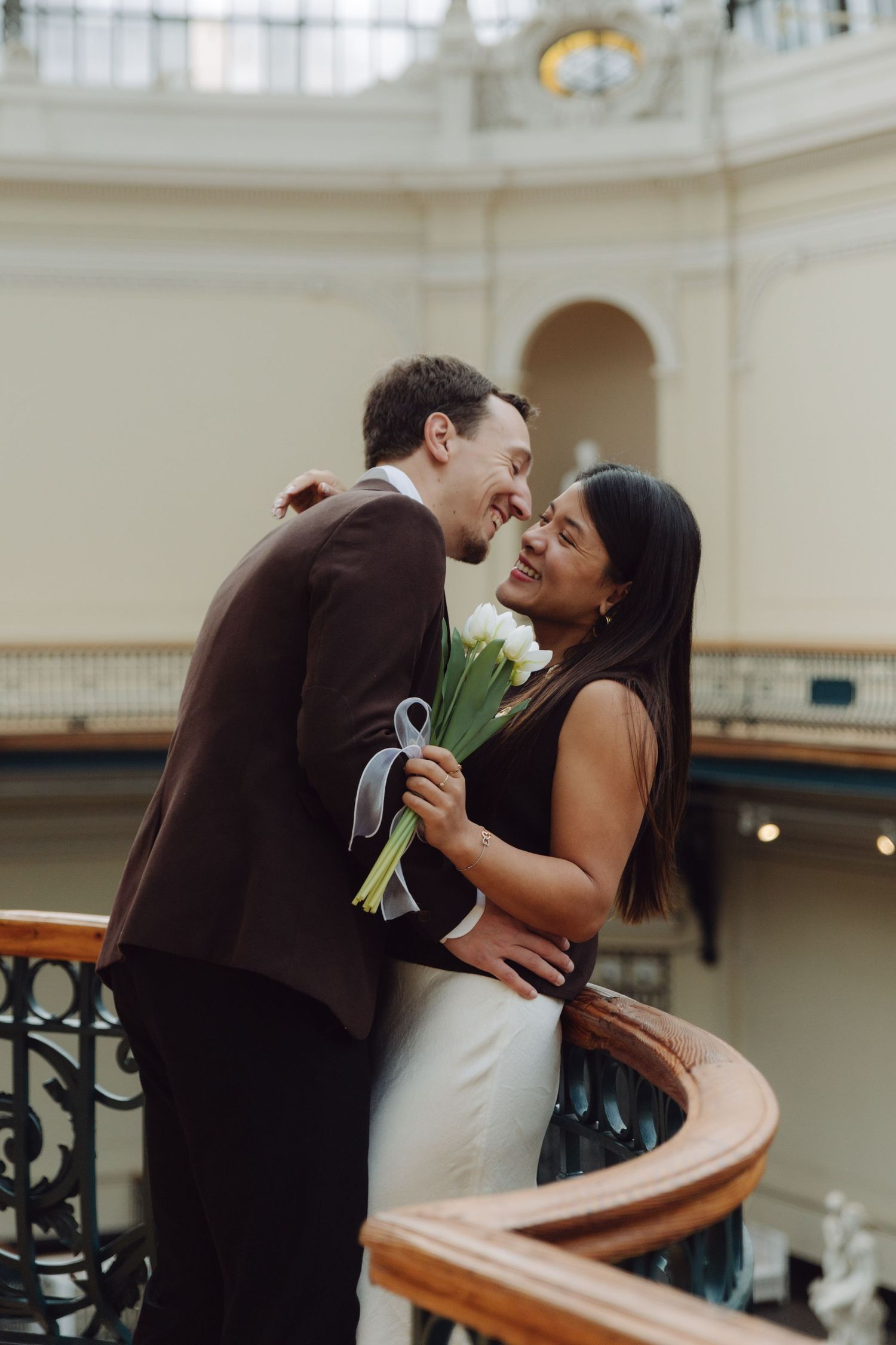 Laughing couple embracing with white tulip bouquet on elegant curved staircase inside ornate historic building.
