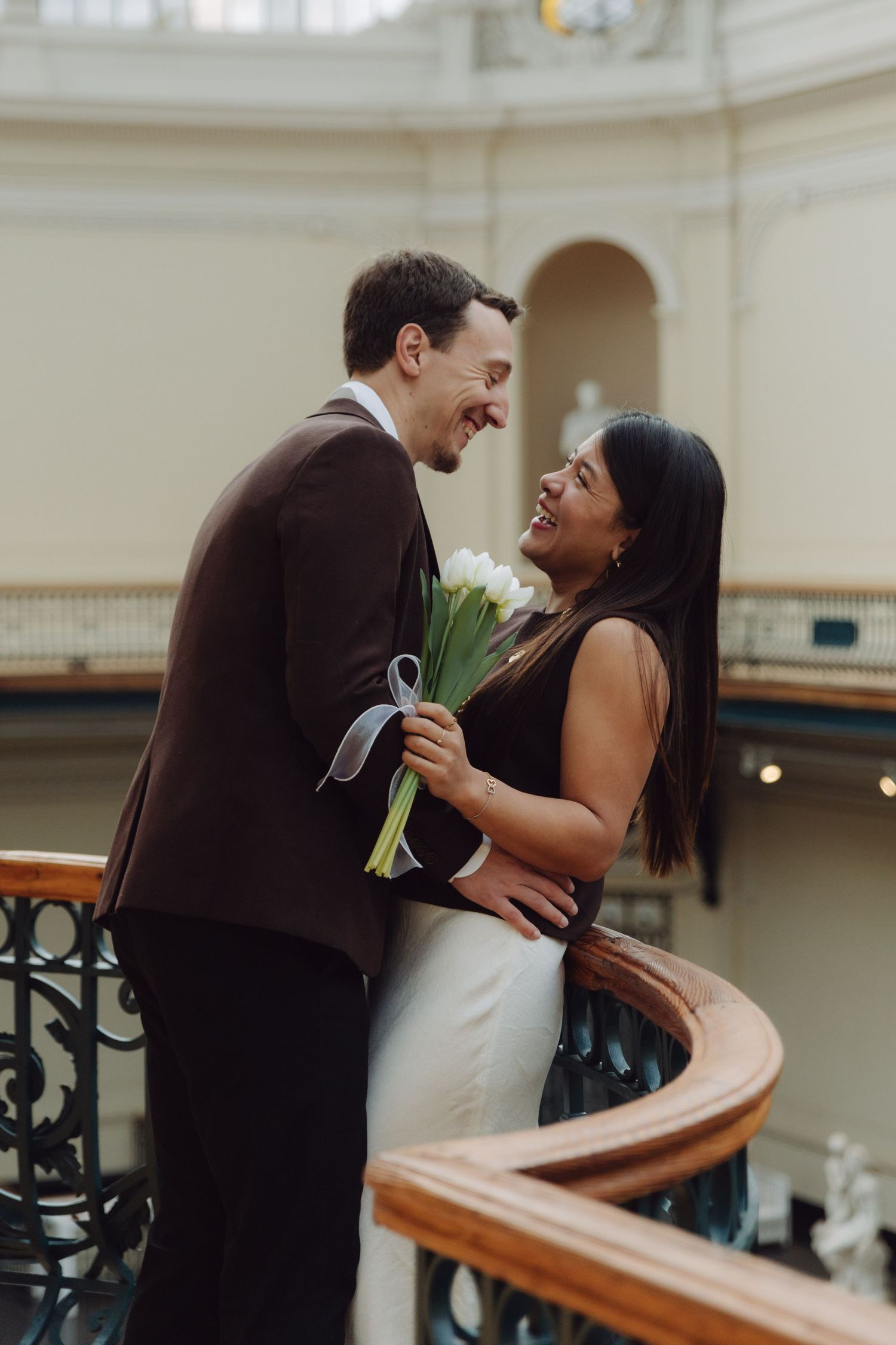 Joyful couple laughing together holding white tulips on curved wooden staircase inside grand classical building.