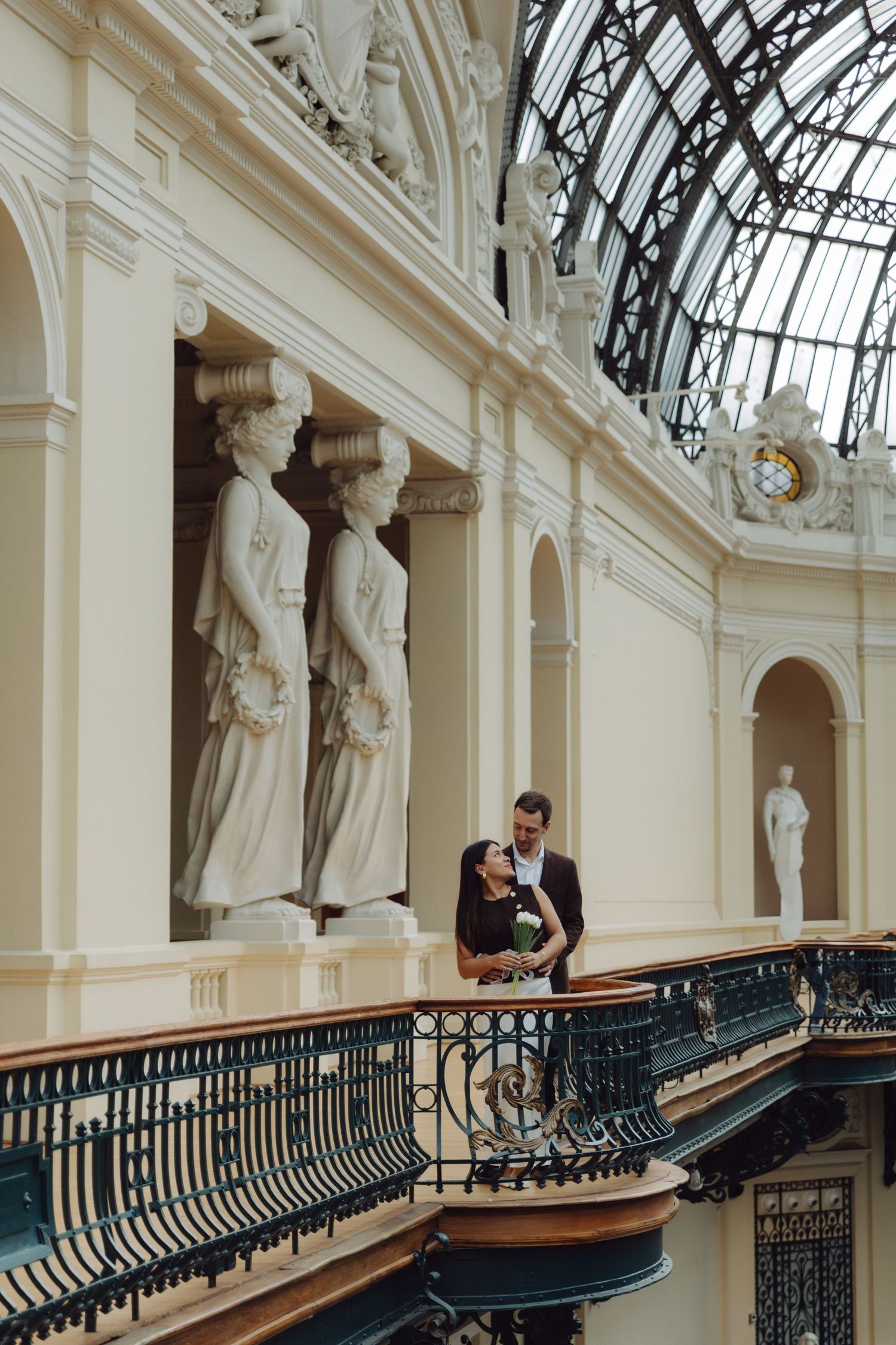 Romantic couple embracing on ornate curved balcony surrounded by classical sculptures inside elegant glass-roofed gallery.