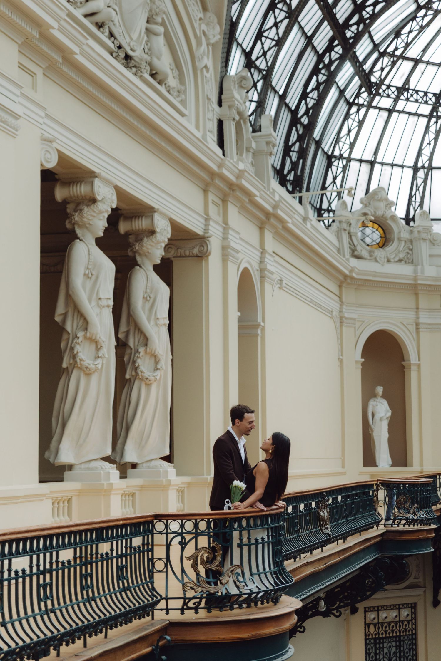 Couple posing romantically on ornate balcony with classical caryatid sculptures in elegant neoclassical building.