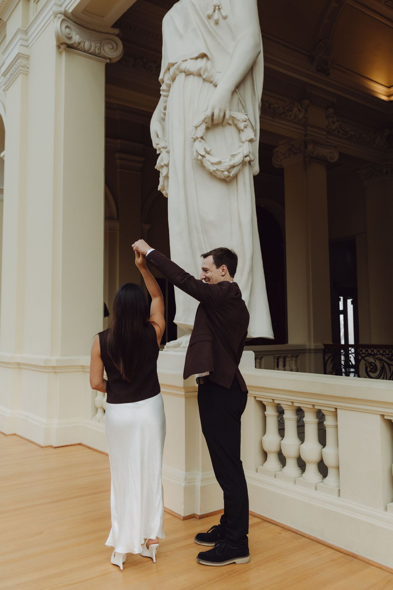 Groom twirling bride in white dress near marble statue on gallery balcony of historic building.