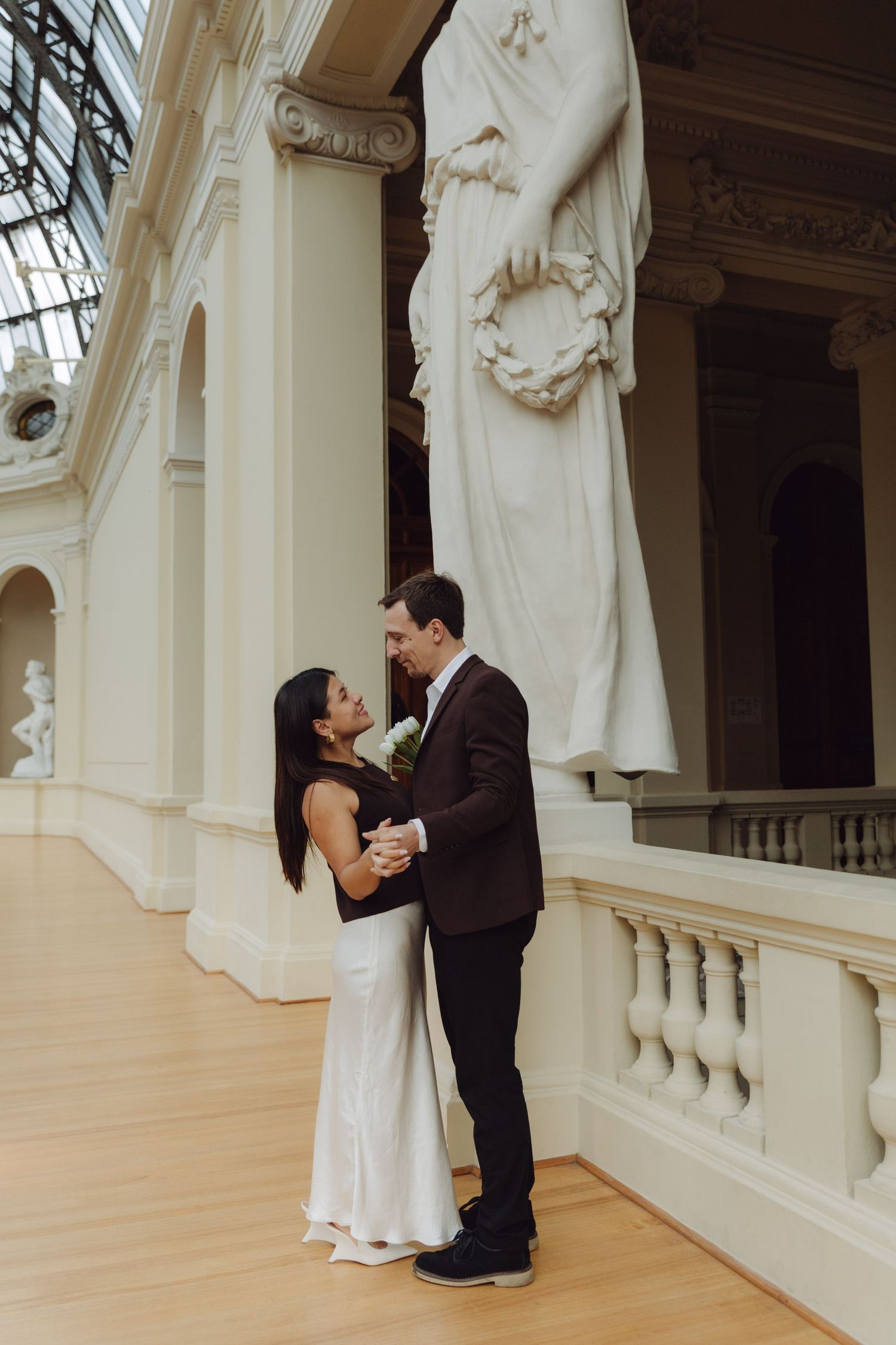 Bride and groom embracing tenderly beside large classical marble statue on museum balcony.