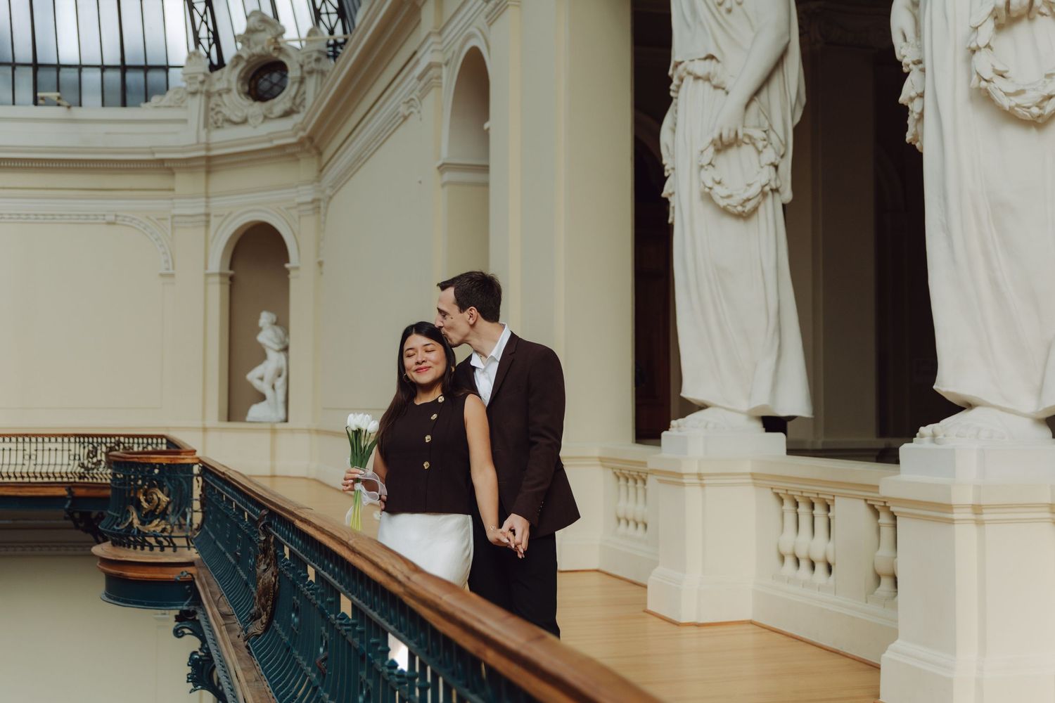 Smiling couple sharing romantic moment on balcony surrounded by classical statues in grand hall.