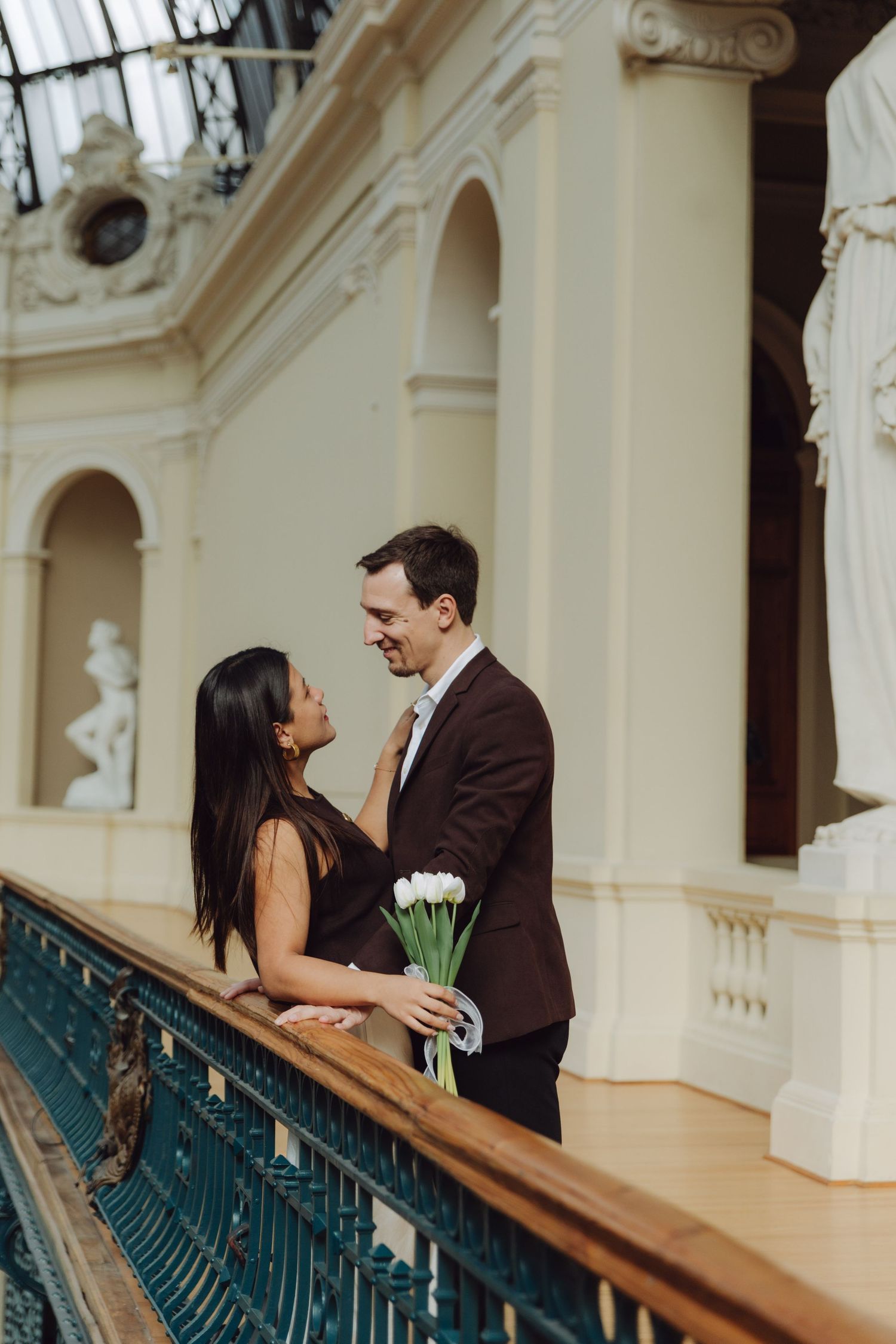Couple gazing lovingly at each other on ornate balcony, groom holding white tulip bouquet.