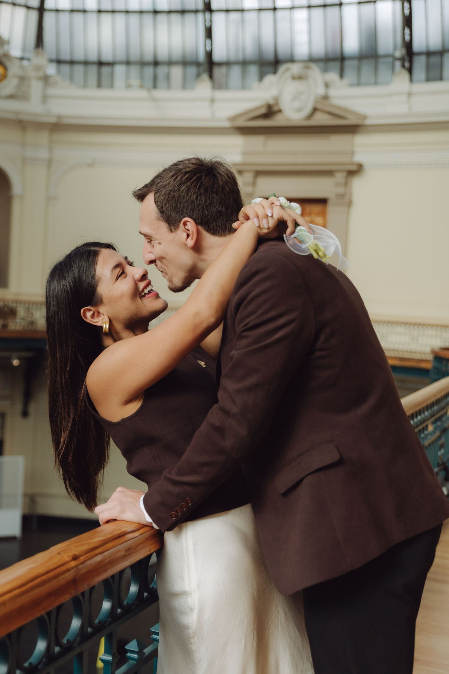 Couple laughing and embracing against wooden railing under glass ceiling of elegant historic venue.