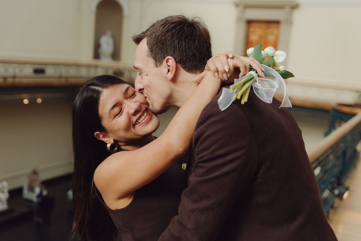 Joyful couple hugging and laughing together, bride holding white tulip bouquet in grand building.