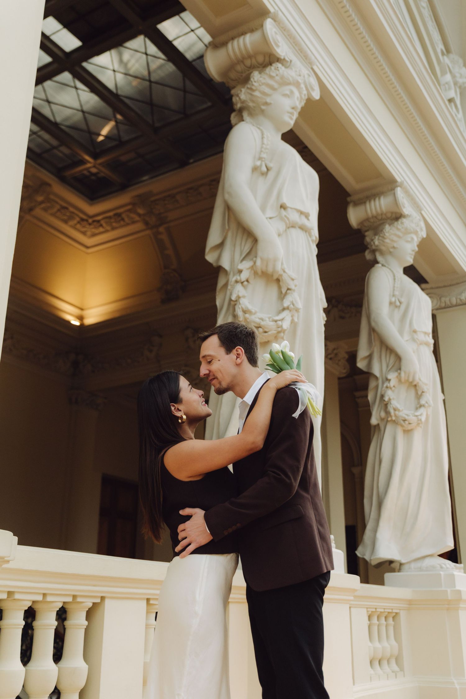 Couple sharing intimate moment near classical caryatid sculptures on balcony of ornate historic building.