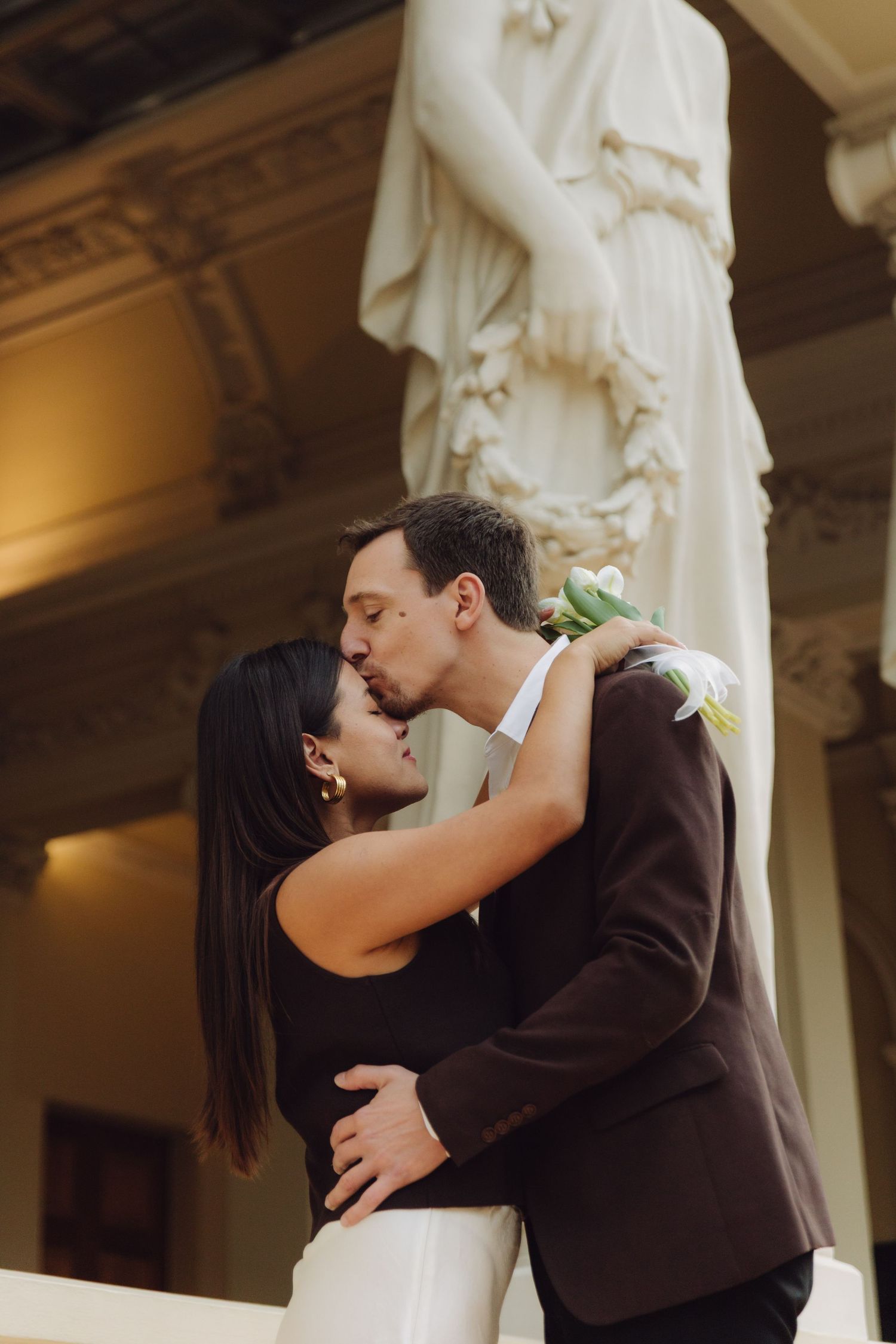 Couple sharing passionate kiss beneath large marble statue on balcony of elegant classical building.
