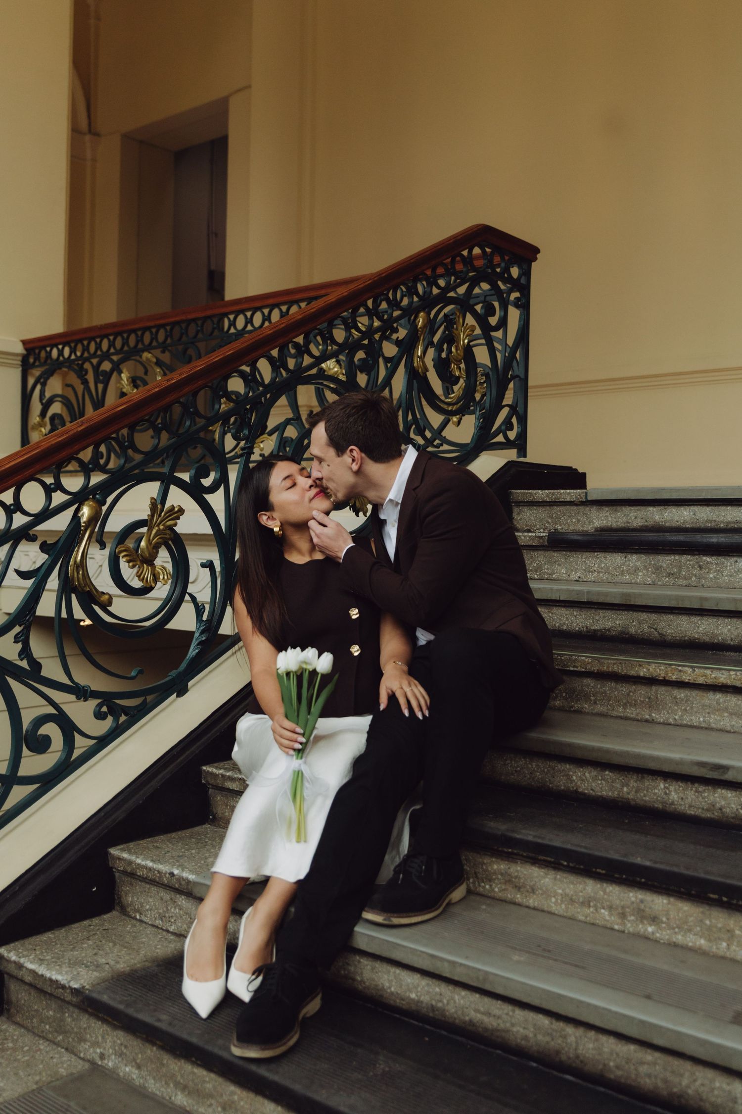 Couple sitting closely on ornate staircase, bride holding white tulips, sharing a tender kiss.