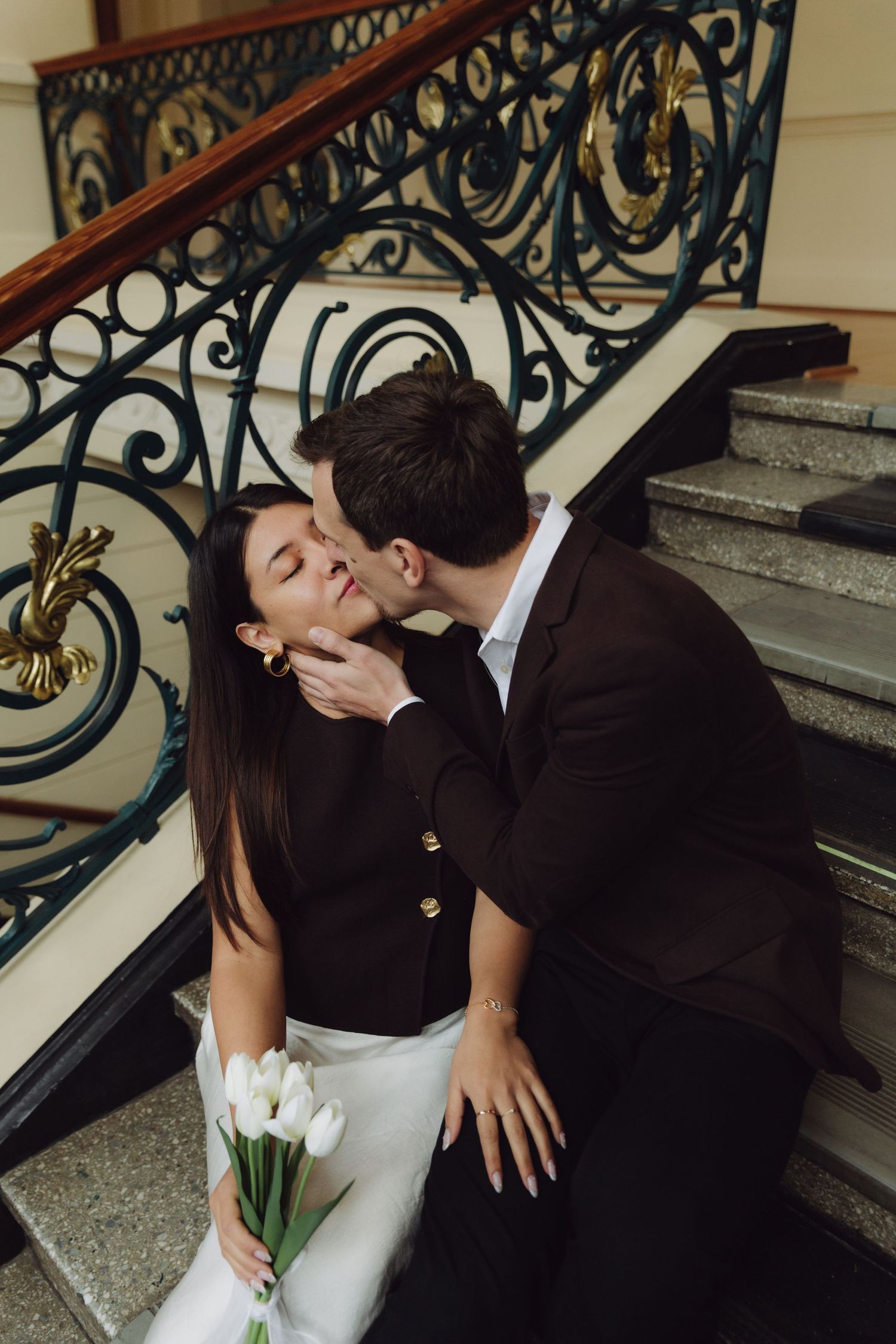 Couple sharing a romantic kiss on ornate wrought iron staircase, woman holding white tulips.