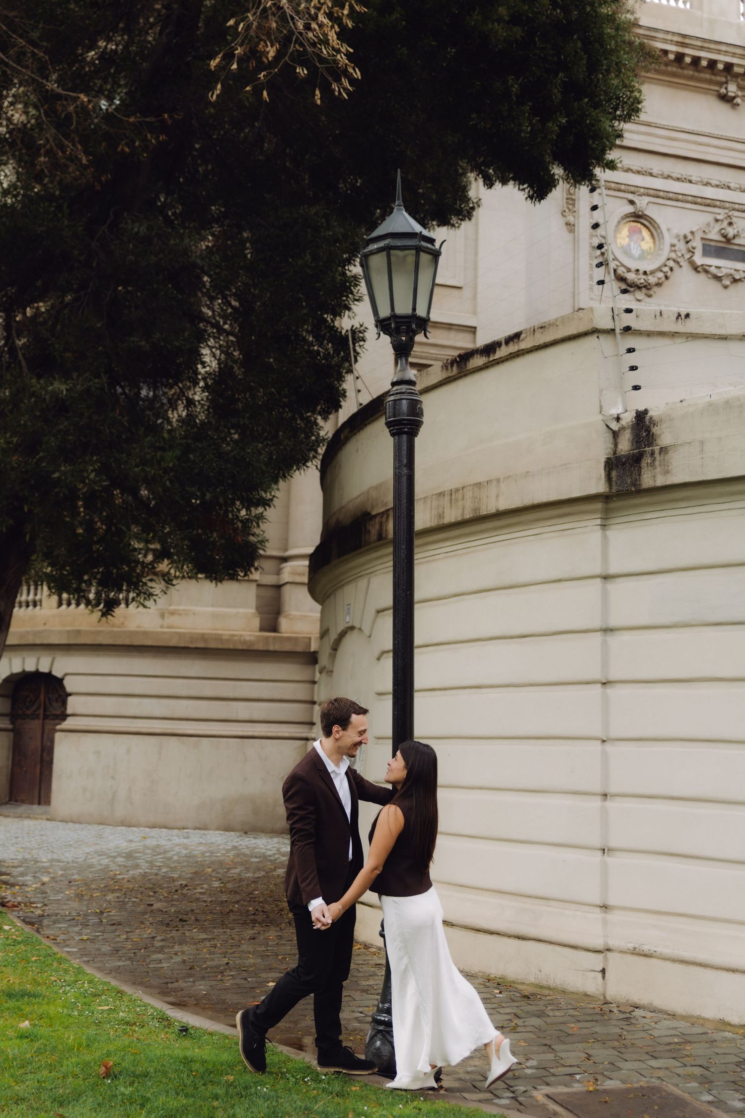 Couple holding hands and smiling near vintage street lamp by neoclassical building exterior.