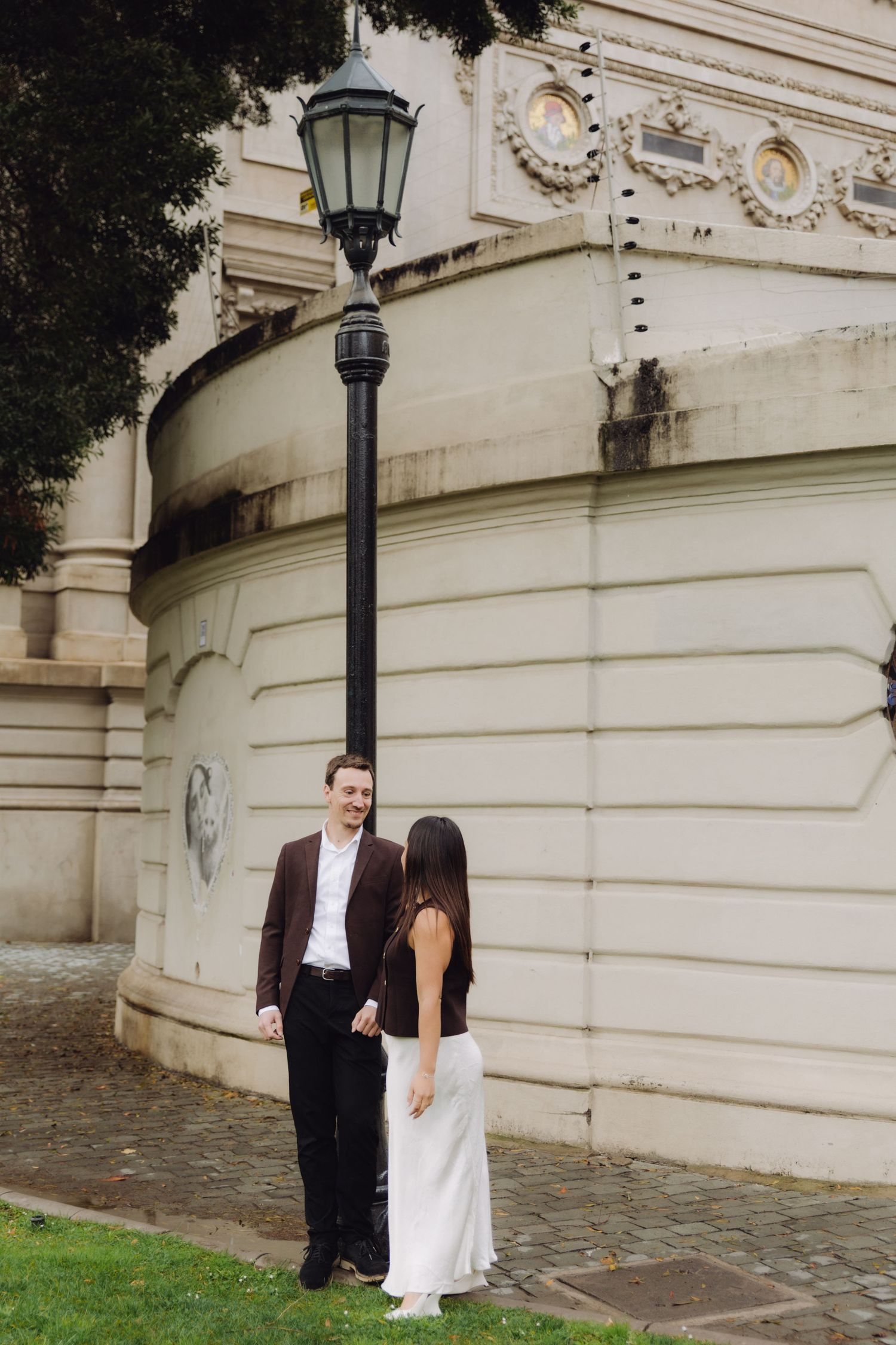 Couple holding hands near ornate street lamp beside grand neoclassical building facade.