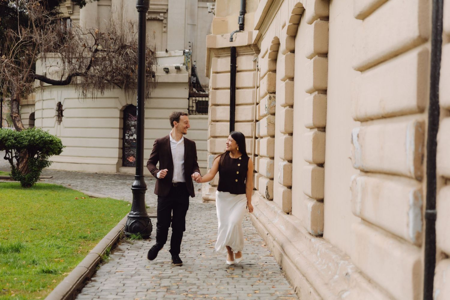 Couple walking hand in hand on cobblestone path beside neoclassical building exterior.