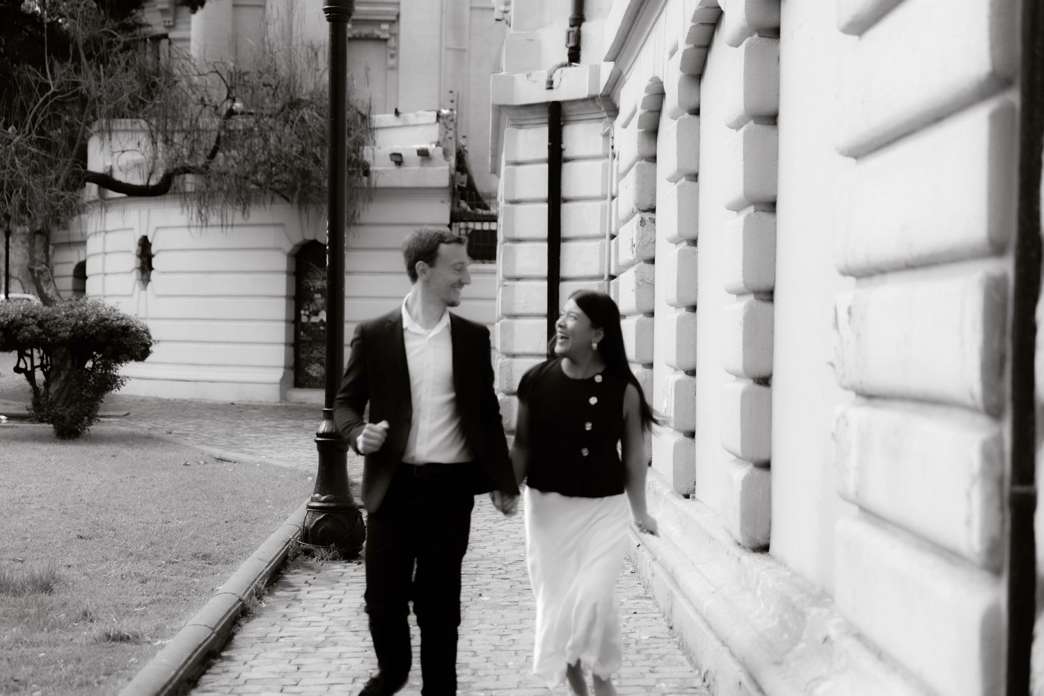Black and white photo of couple walking hand in hand along cobblestone street.