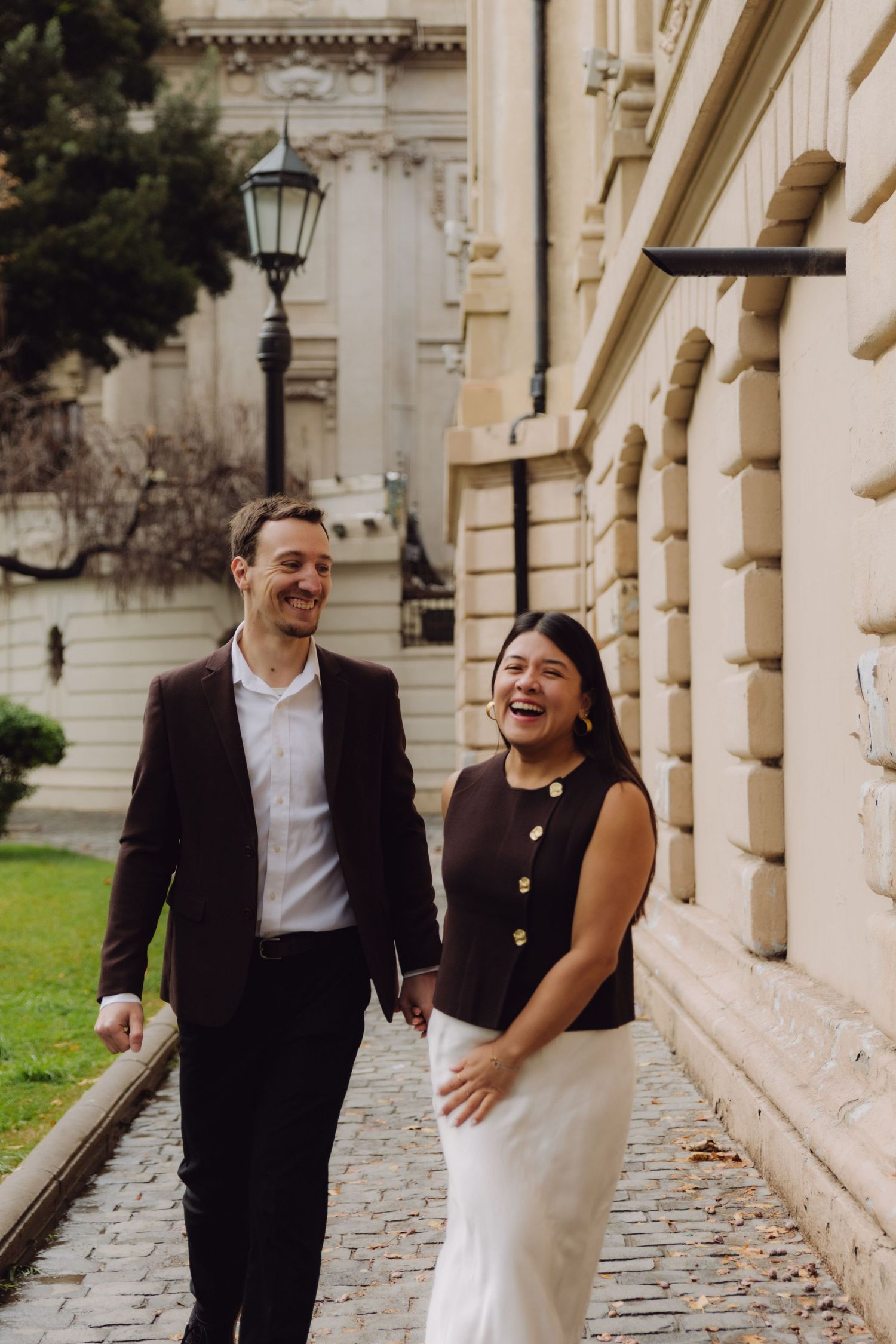 Laughing couple holding hands while strolling along cobblestone path by historic building.