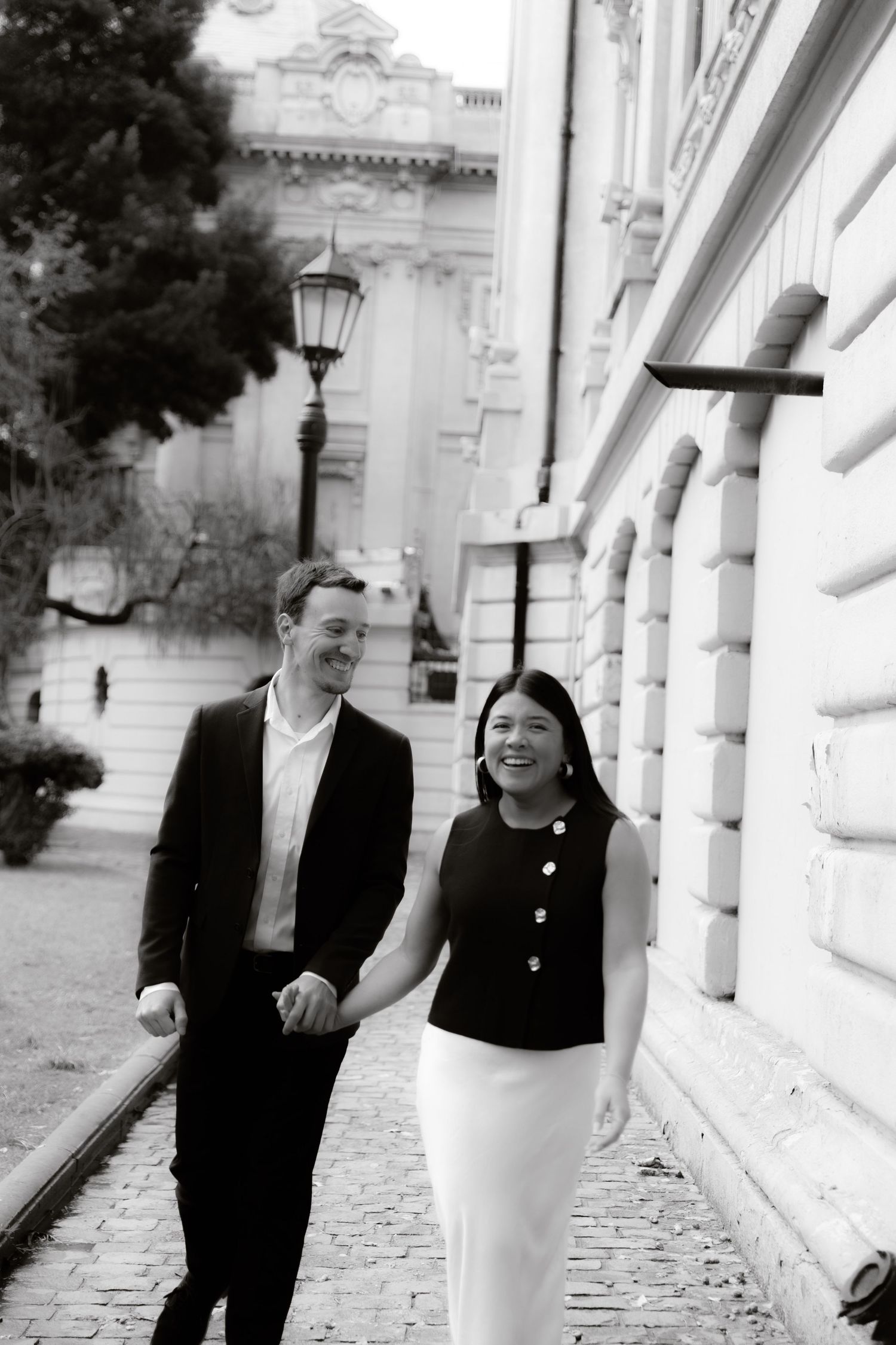 Black and white photo of smiling couple holding hands walking beside historic building.