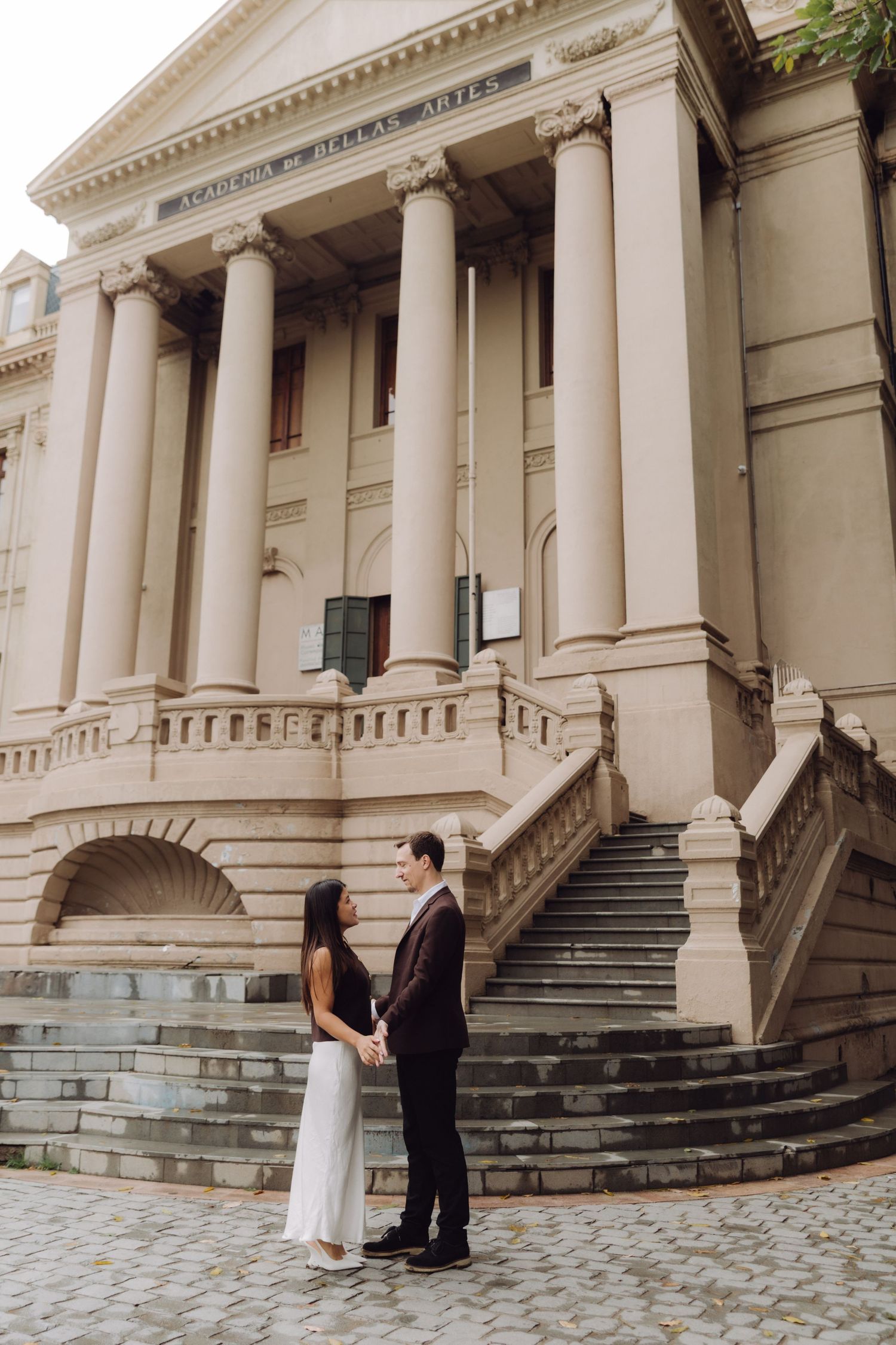 Couple holding hands facing each other before grand neoclassical Academia Bellas Artes building.
