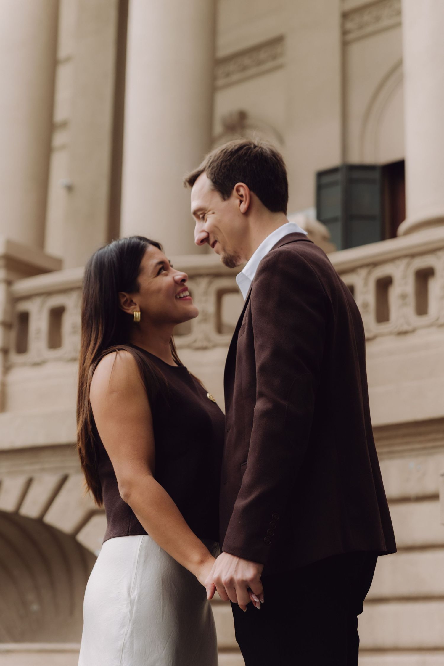 Couple holding hands and smiling at each other in front of a classical building during an engagement session.