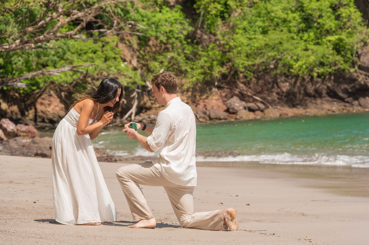 Man proposes on a tropical beach while woman reacts with joy, lush green trees and turquoise water in the background.