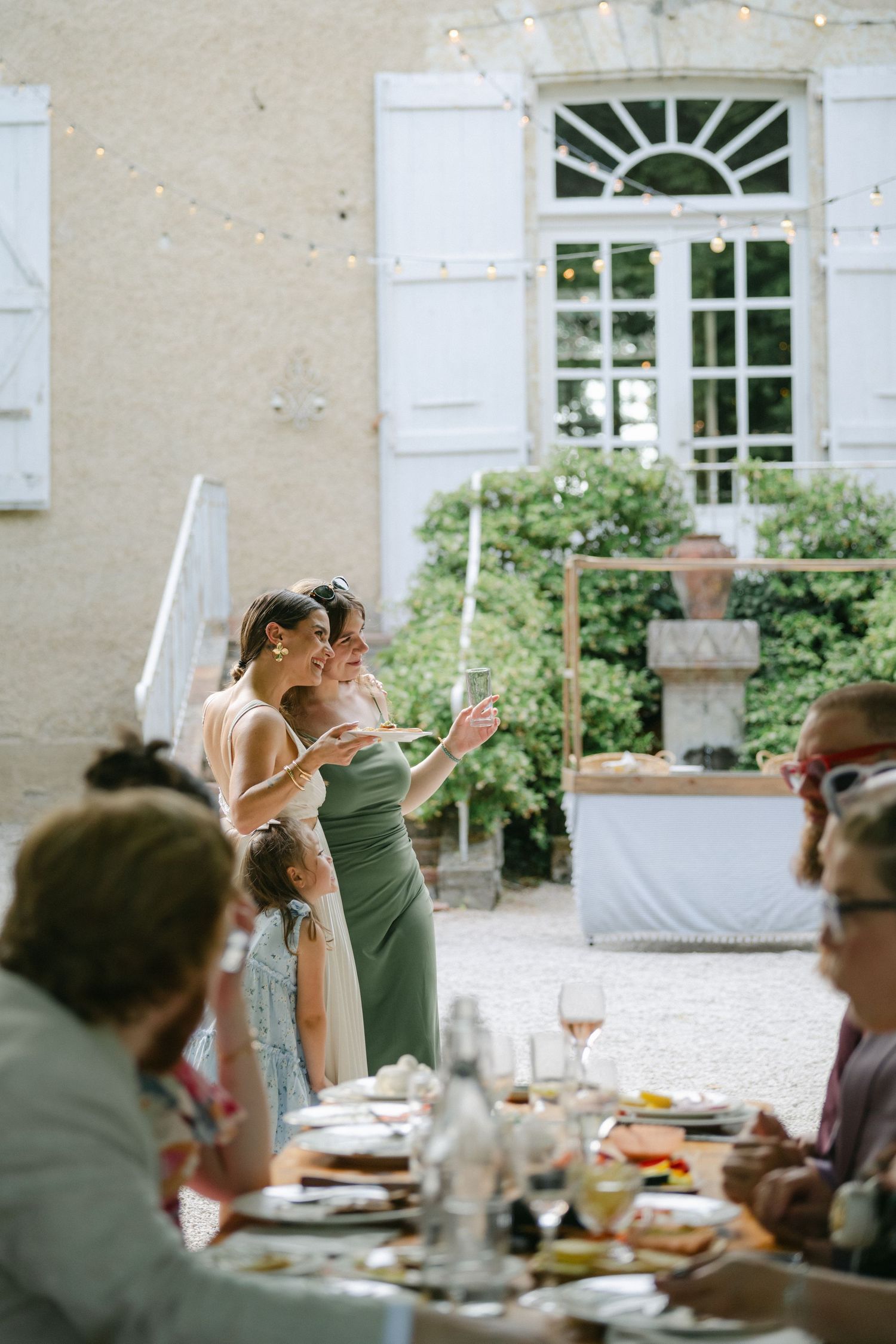 Two bridesmaids in elegant dresses toast at an outdoor wedding reception beside a French chateau.