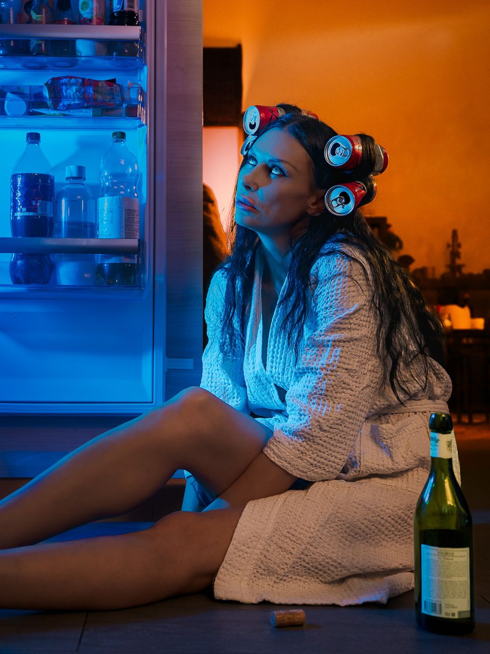 Woman in bathrobe with hair rollers sits on floor by open fridge at night with wine bottle nearby.