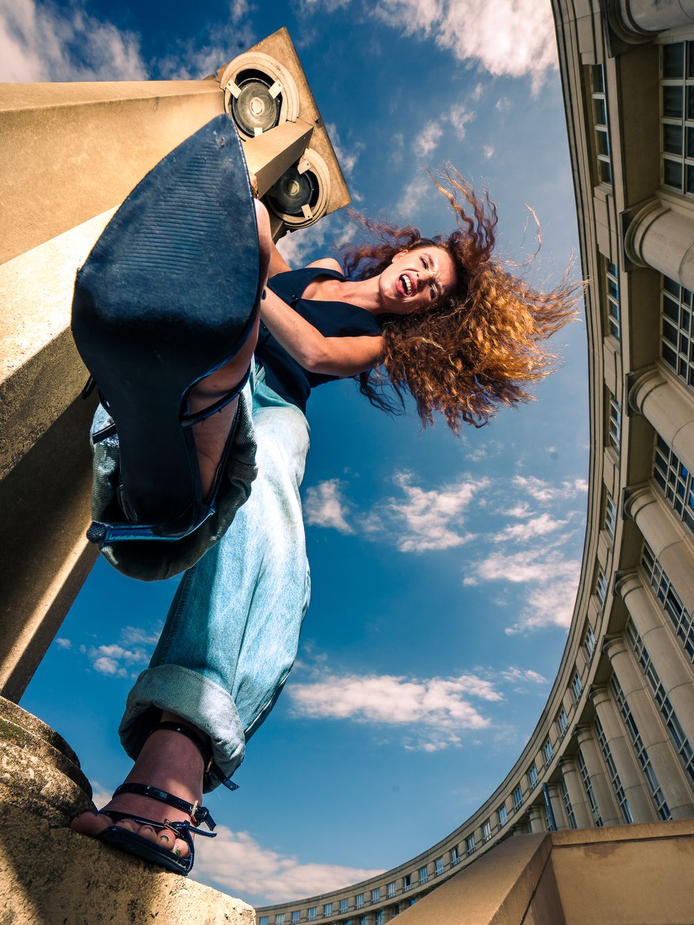 Woman with curly red hair laughing, holding a speaker outdoors, shot from low angle with blue sky background.