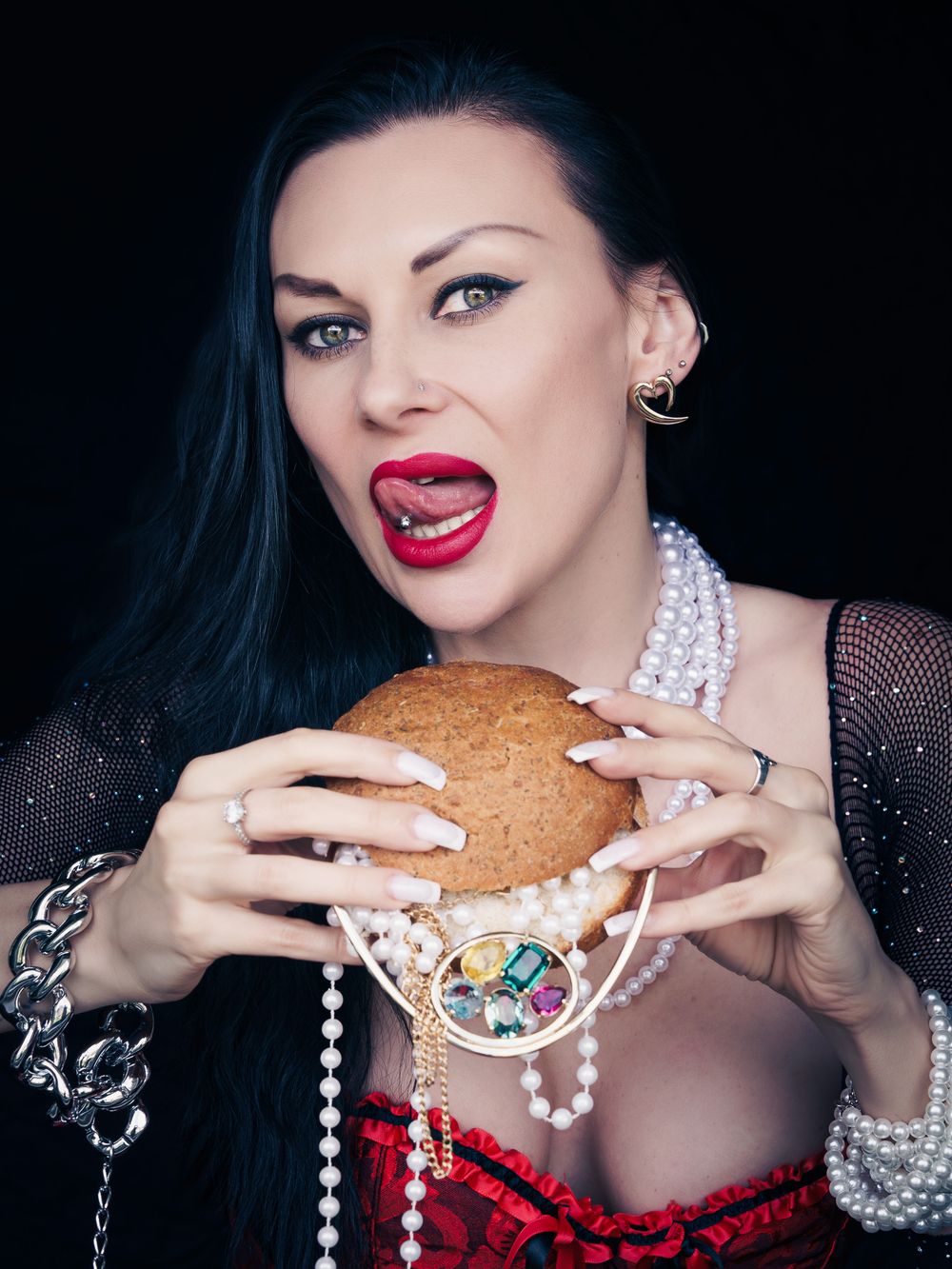 Woman with long nails, pearl necklace, and red corset holding a large burger with mouth open.
